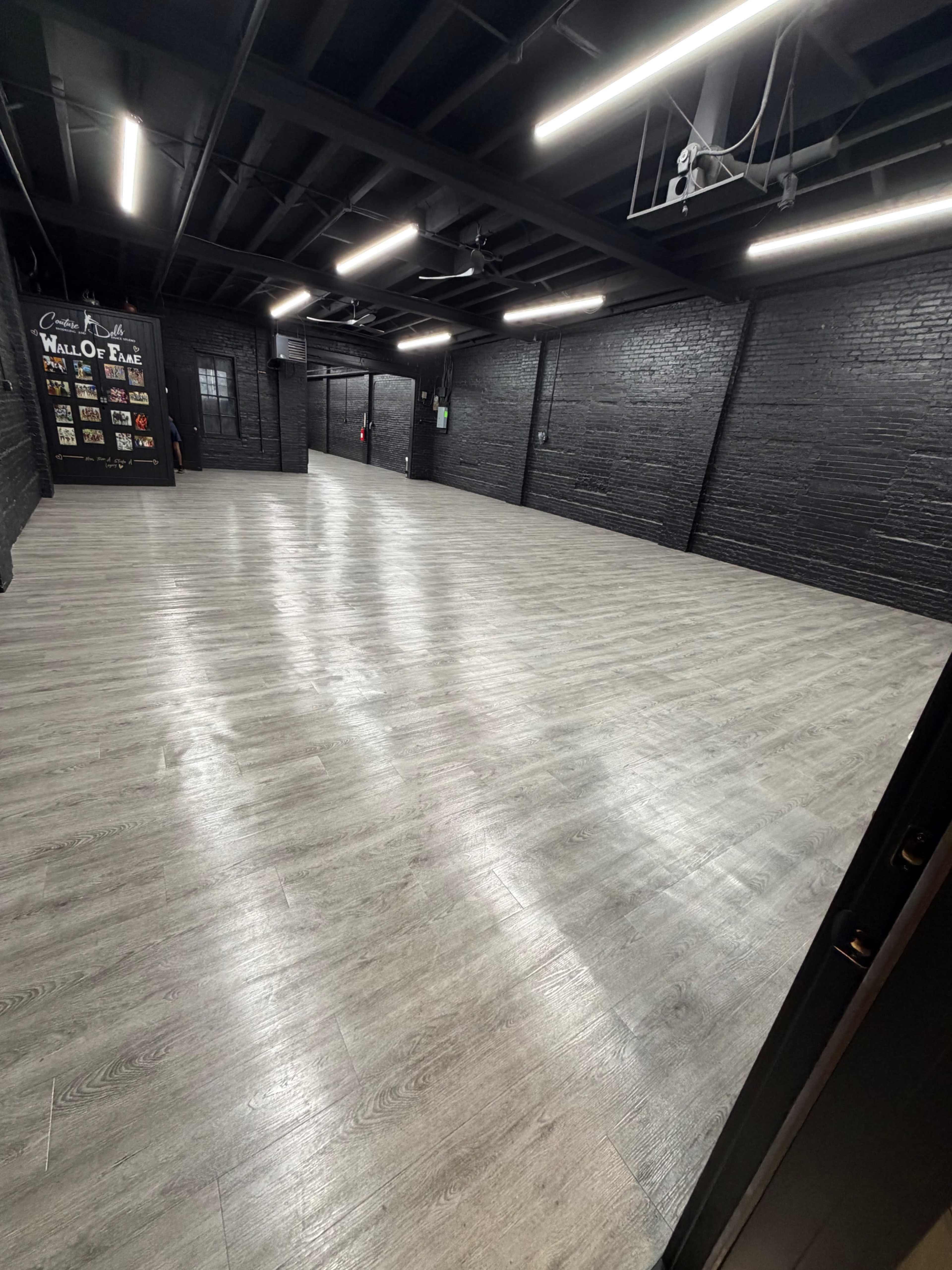 The image shows a spacious, empty room with gray wooden flooring and black brick walls, illuminated by ceiling lights.