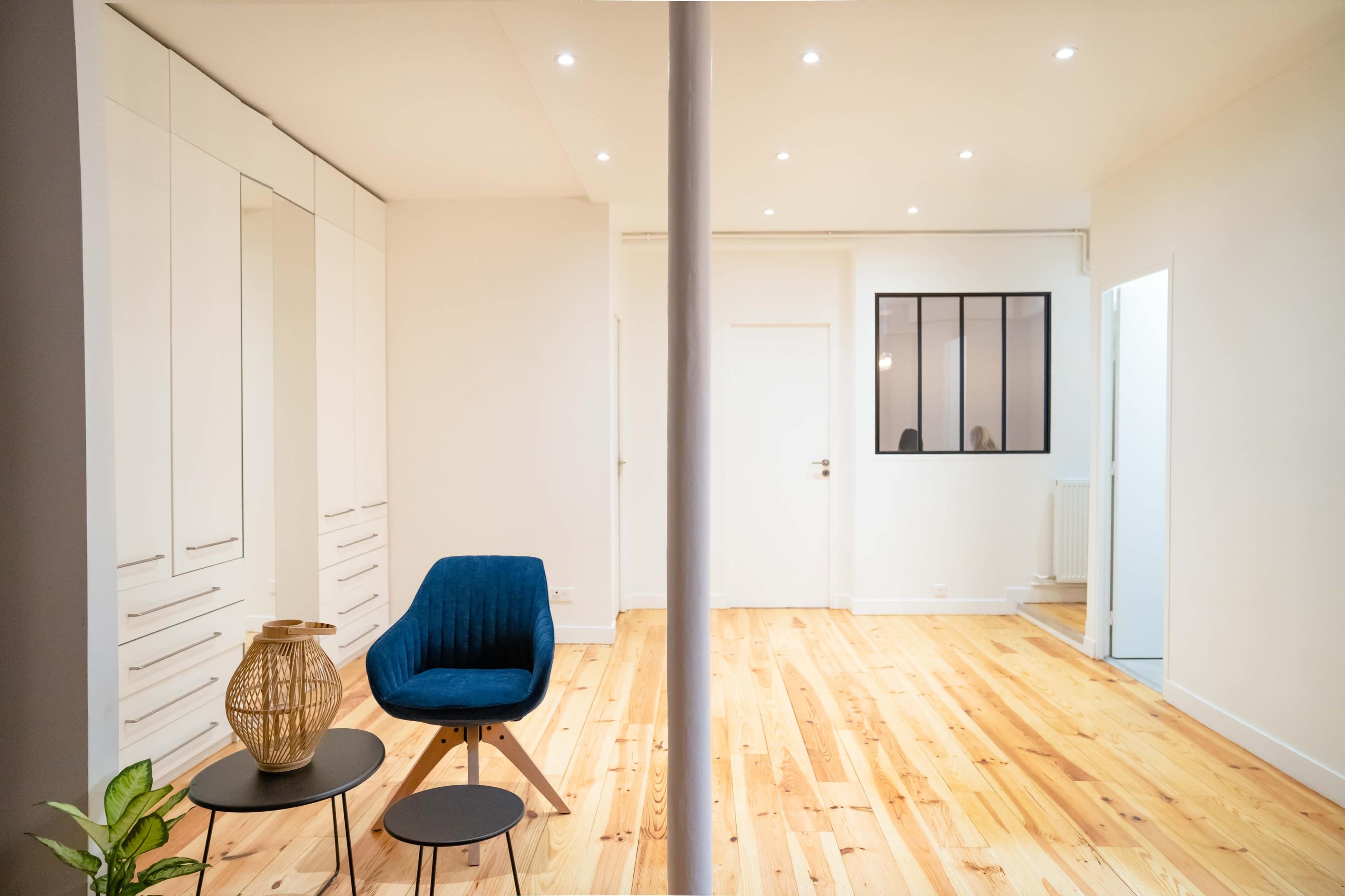 A modern interior features a blue chair and small table in a room with light-colored wooden flooring and white cabinetry.