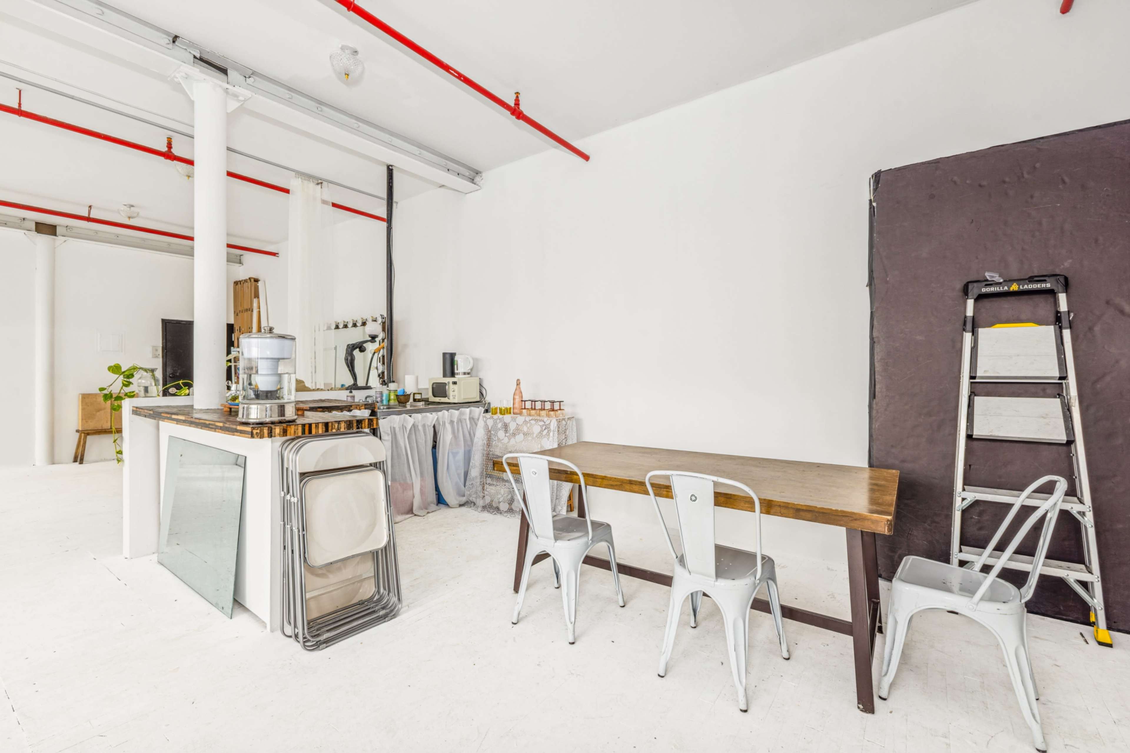 The image shows a spacious, brightly lit kitchen area featuring a wooden table with chairs, a water dispenser, and kitchen equipment against a plain white wall.