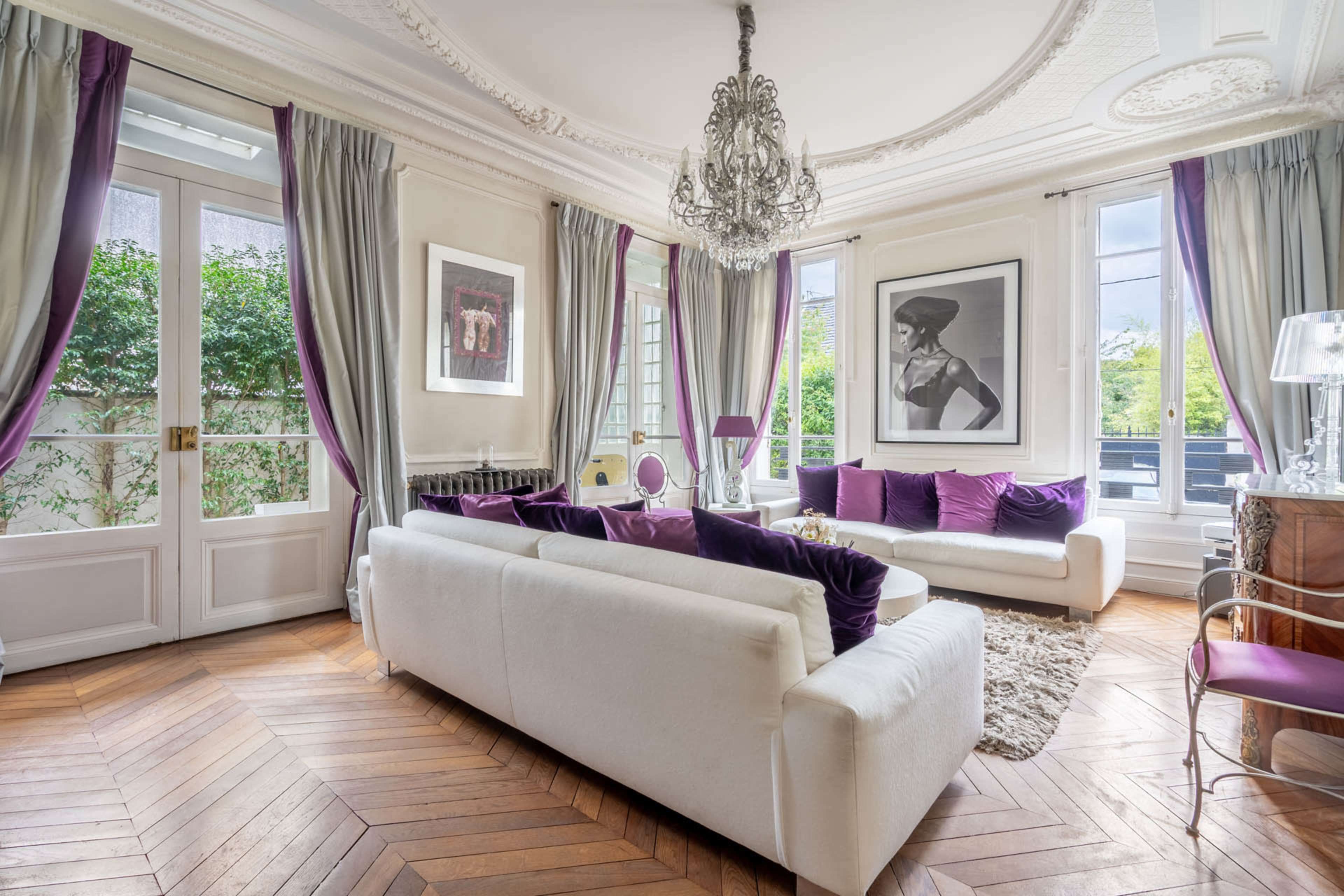 A well-decorated living room features a white sofa, purple accents, a large artwork on the wall, and elegant window treatments.