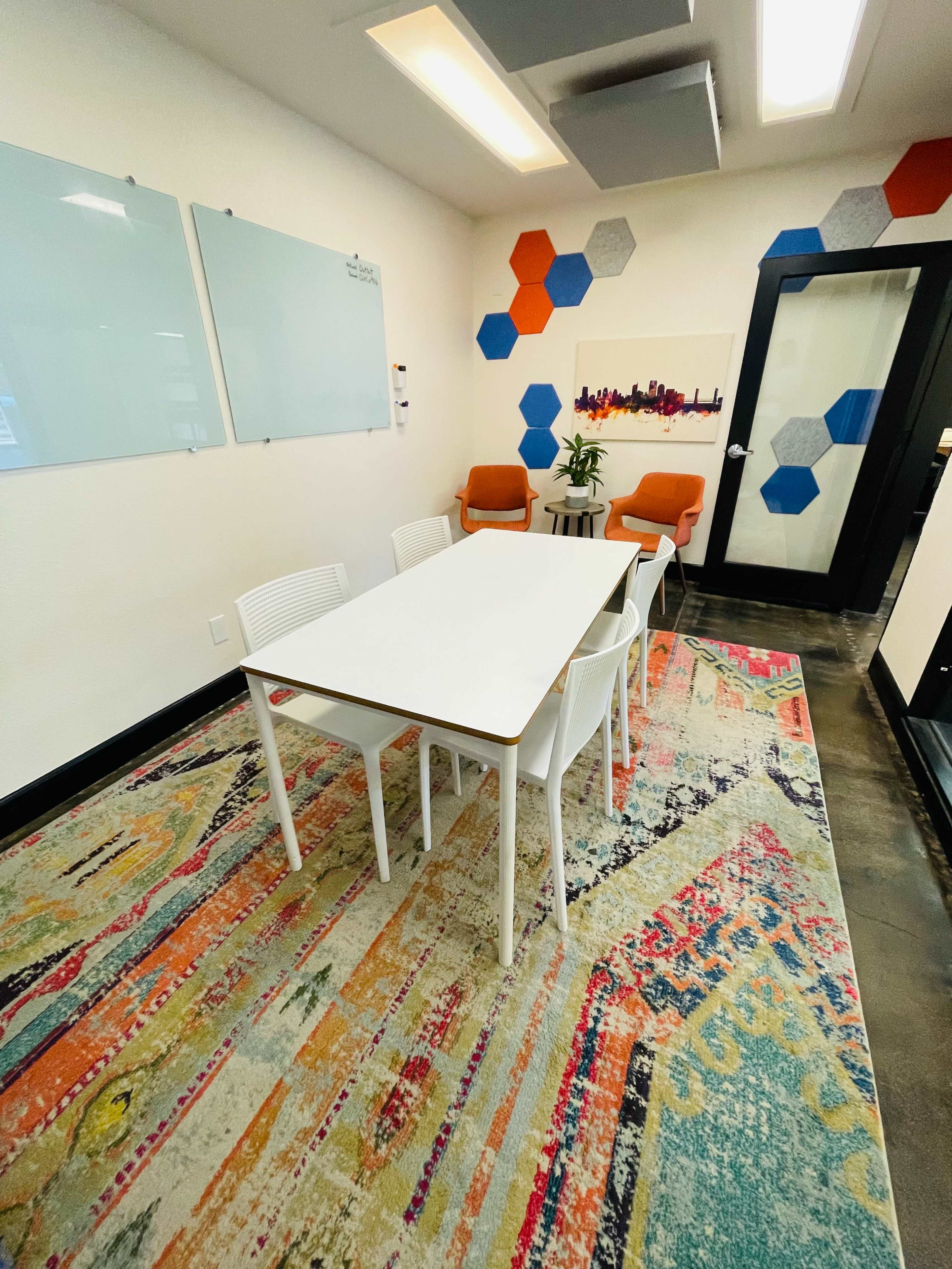 A modern meeting room featuring a white table surrounded by six chairs, colorful rug flooring, and orange accent chairs against a wall with hexagon décor.