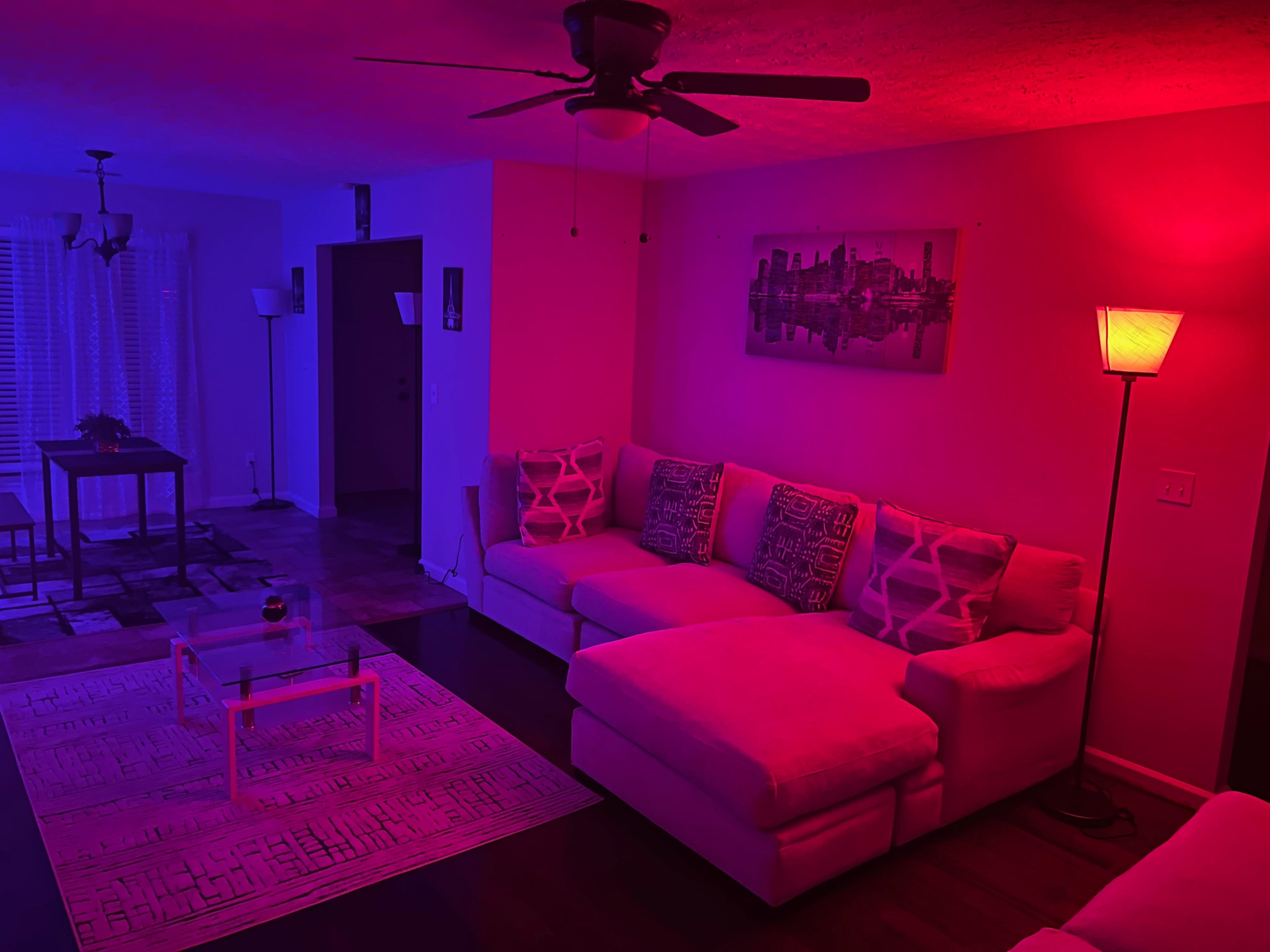 The living room features a sectional sofa, a coffee table, and a dining area, illuminated by colored lighting in shades of red and blue.