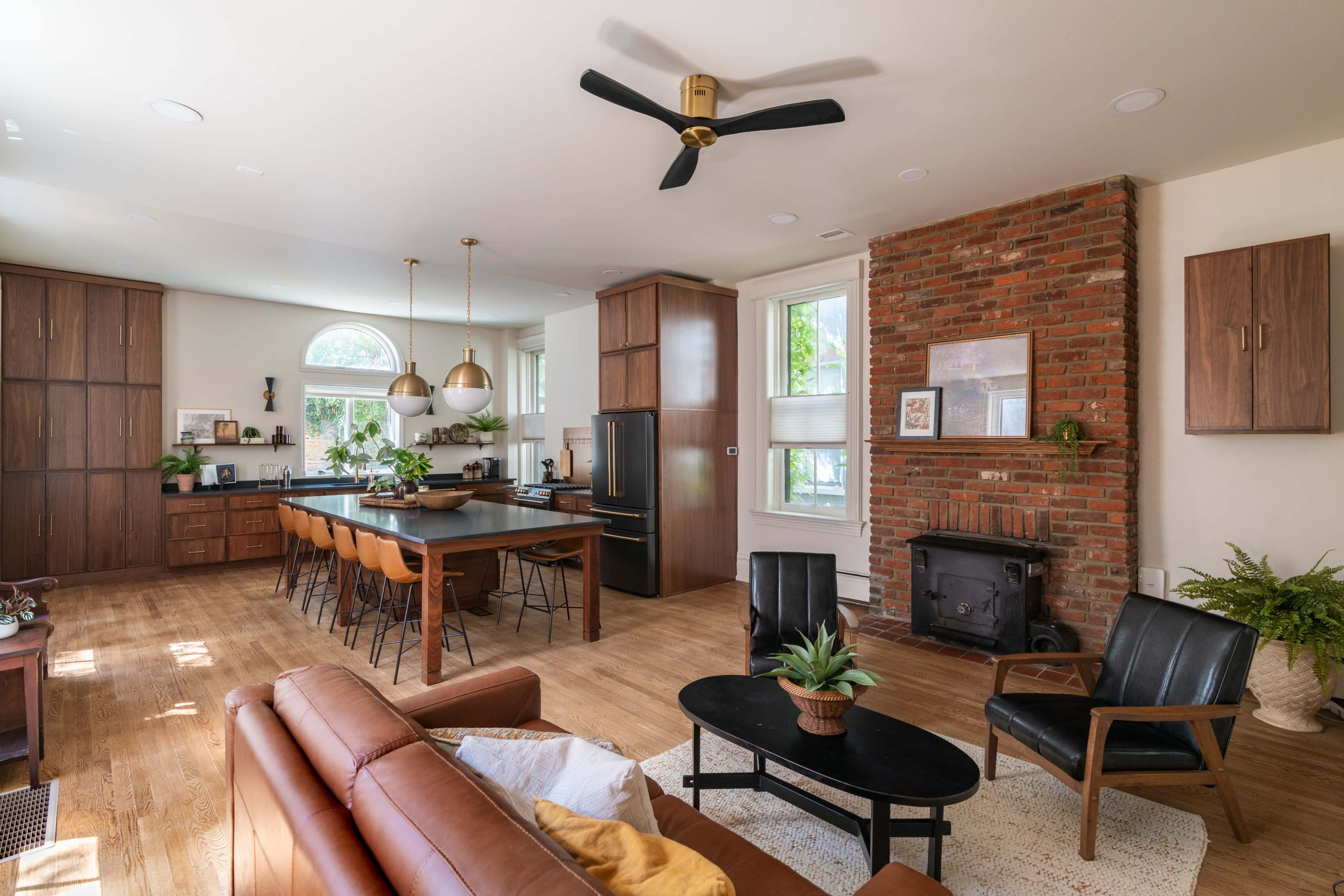 The image shows a modern kitchen and living area featuring a brick fireplace, wooden cabinetry, and a large dining table with leather chairs.