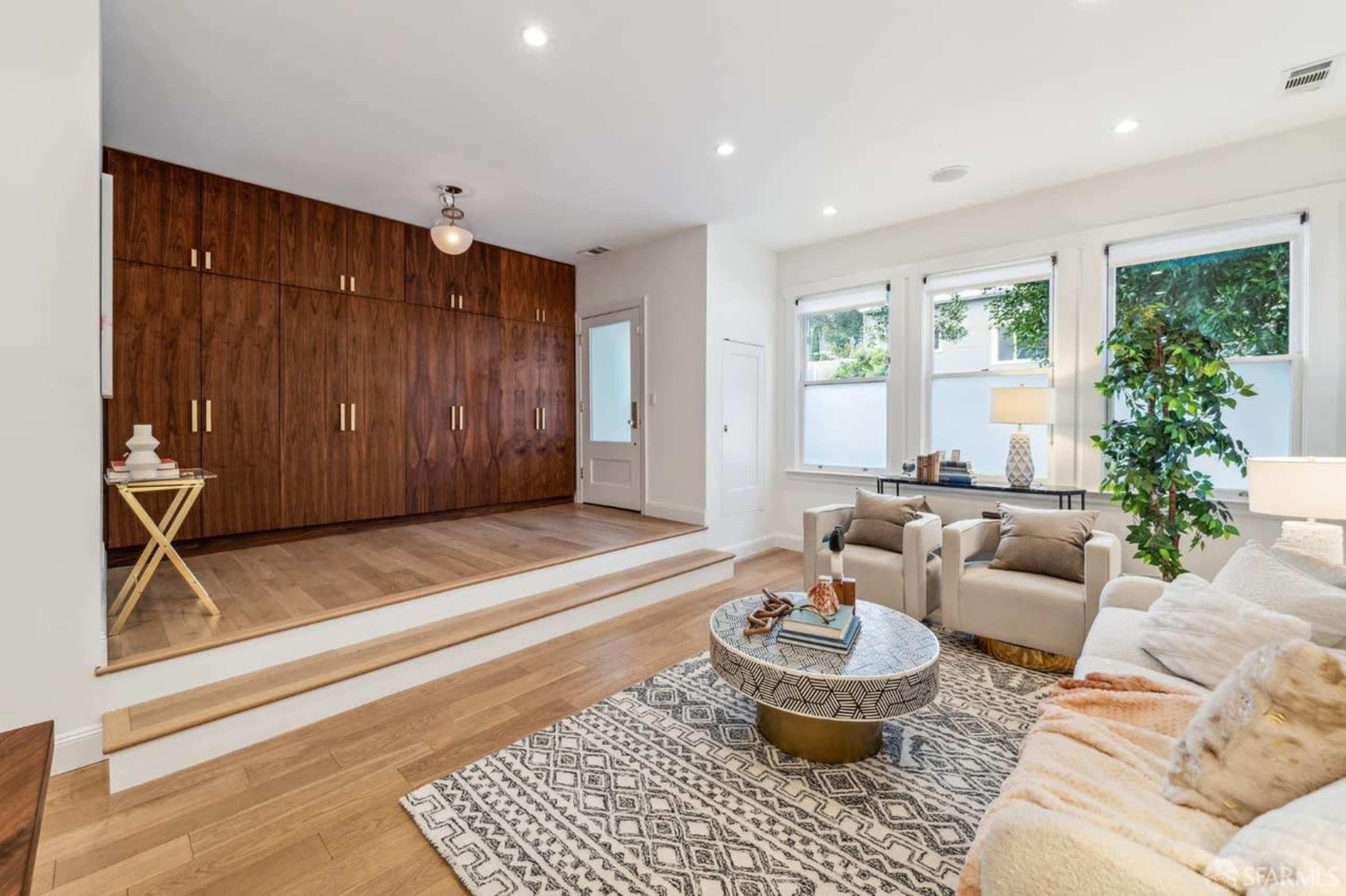 A spacious living room with light wood flooring, a seating area, and a wall of built-in wooden cabinets.