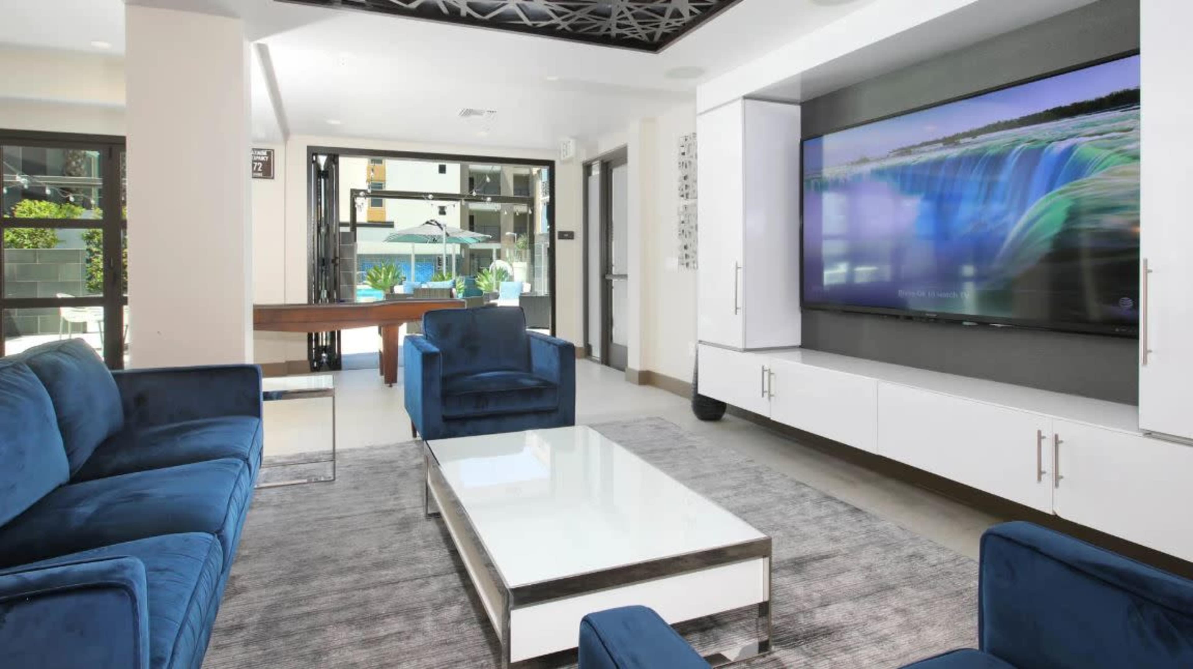 A contemporary living room with blue upholstered sofas, a glass coffee table, and a large flat-screen TV mounted on the wall.