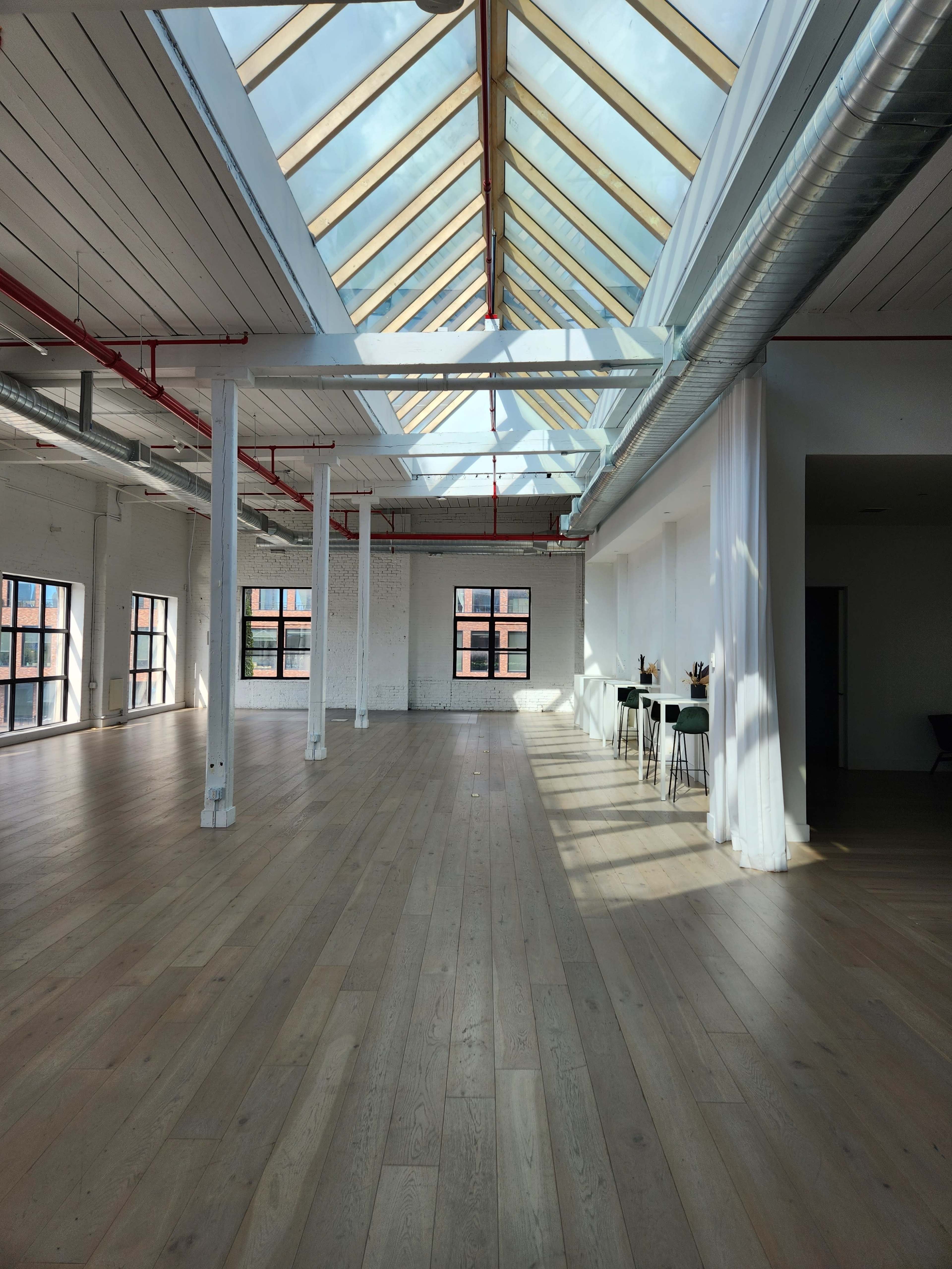 The image shows a spacious interior with wooden floors, large windows, and a skylight that lets in natural light, complemented by minimalist furnishings at one end.