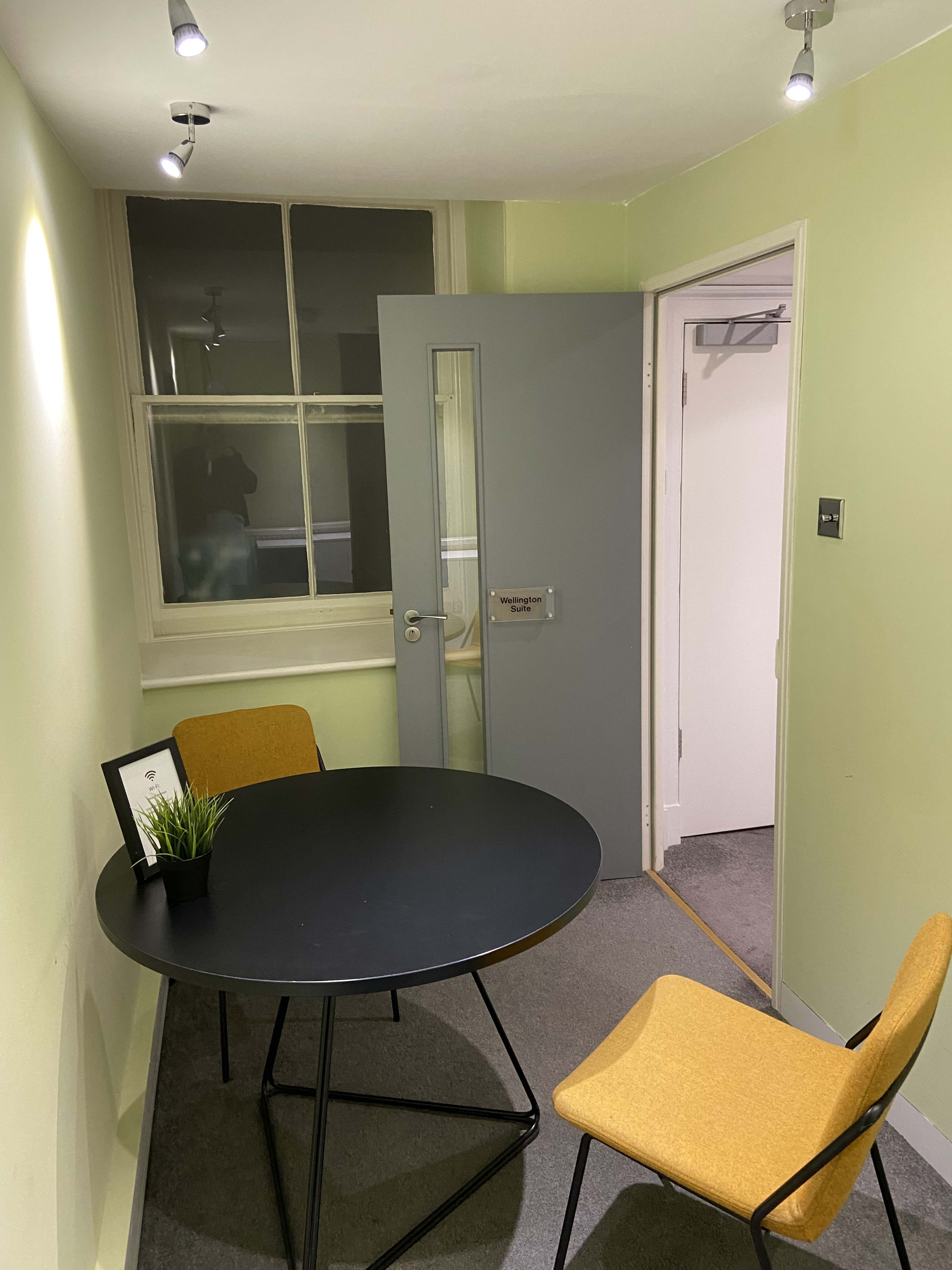 A small room with a round black table and two mustard-colored chairs, featuring a door partially open to an adjacent space.