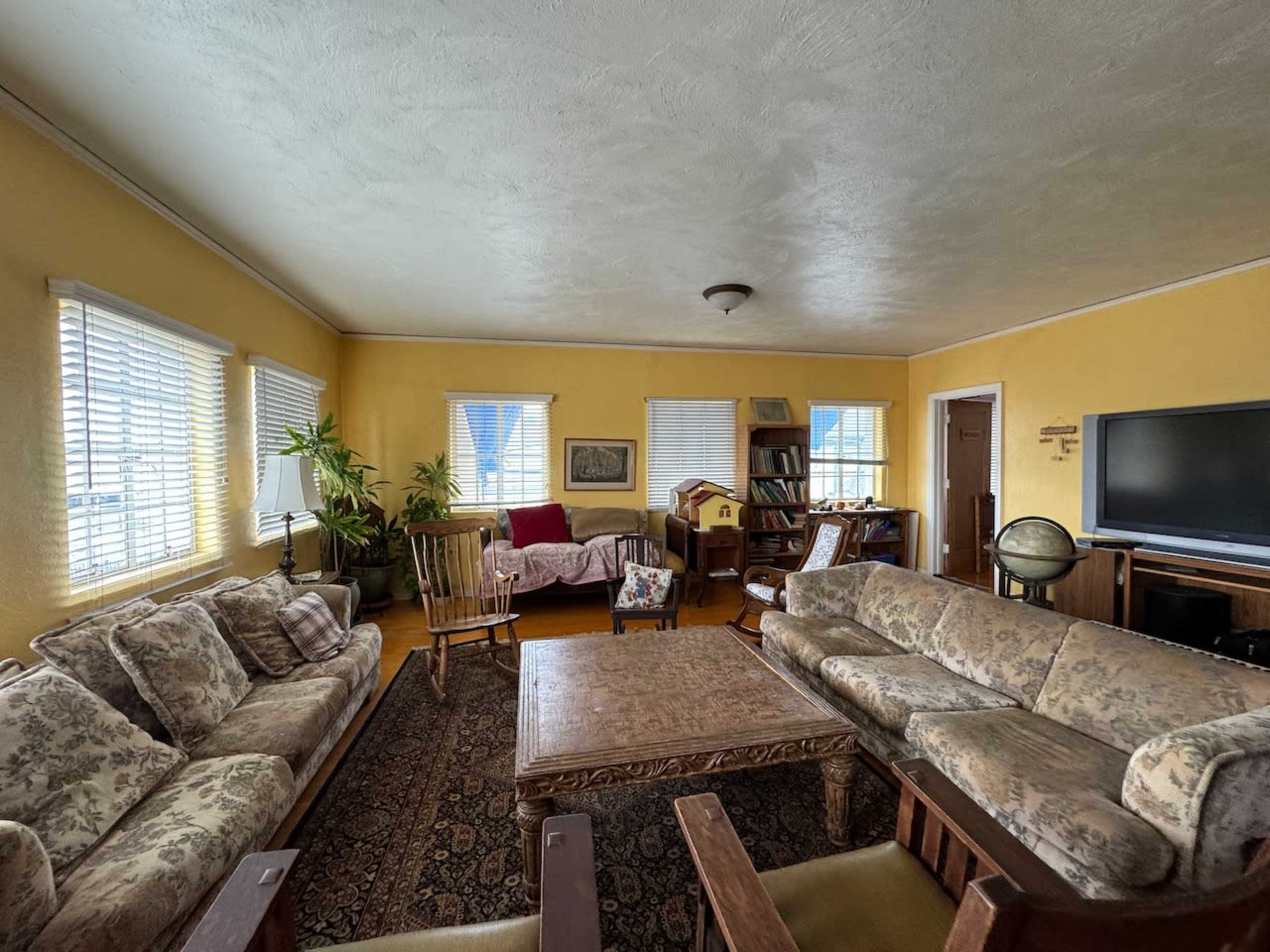 The living room features yellow walls, several sofas, a wooden coffee table, and a variety of plants and furniture surrounding a television.