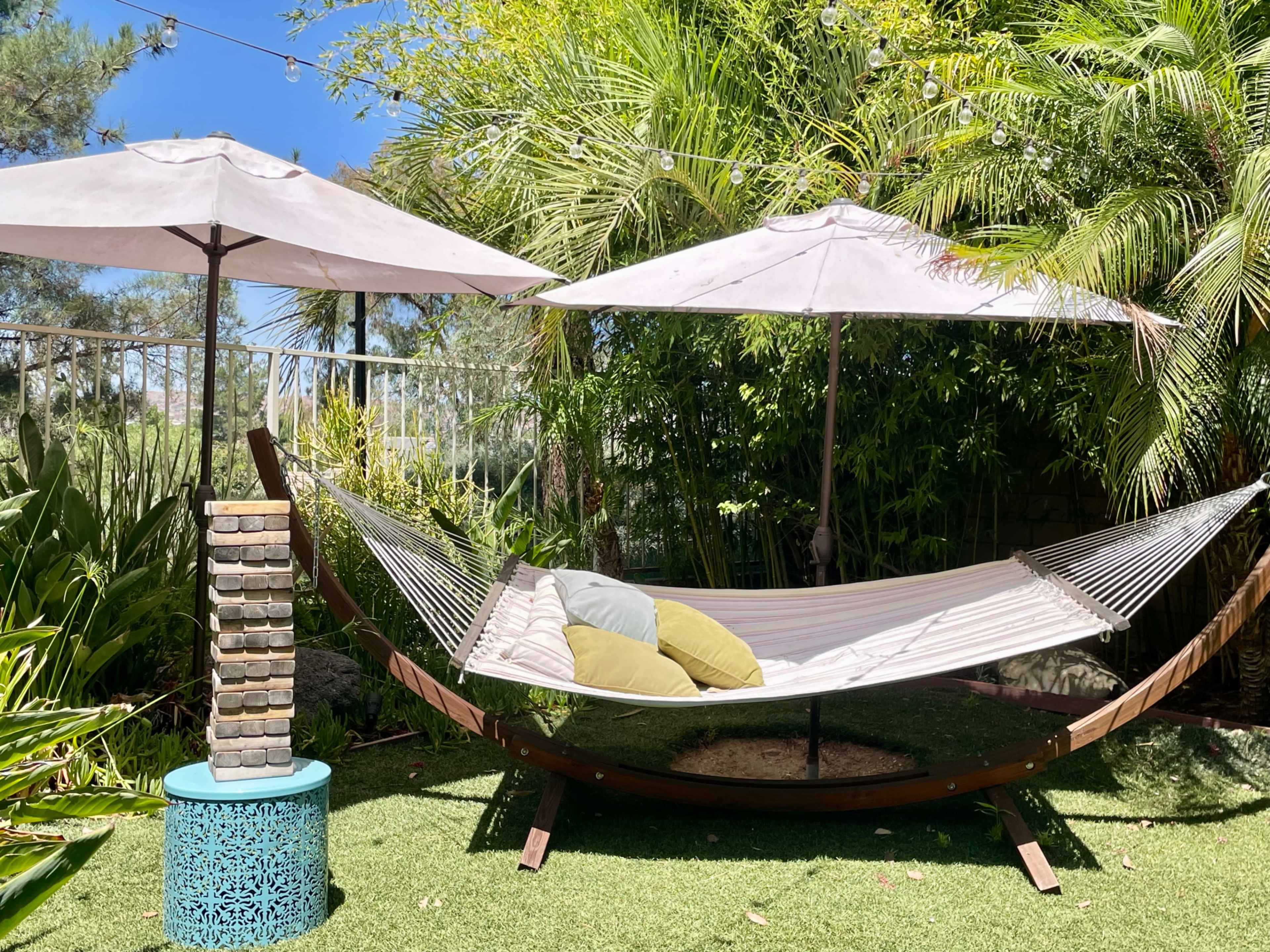 A hammock with green pillows is suspended between two umbrella-topped supports in a lush garden setting.