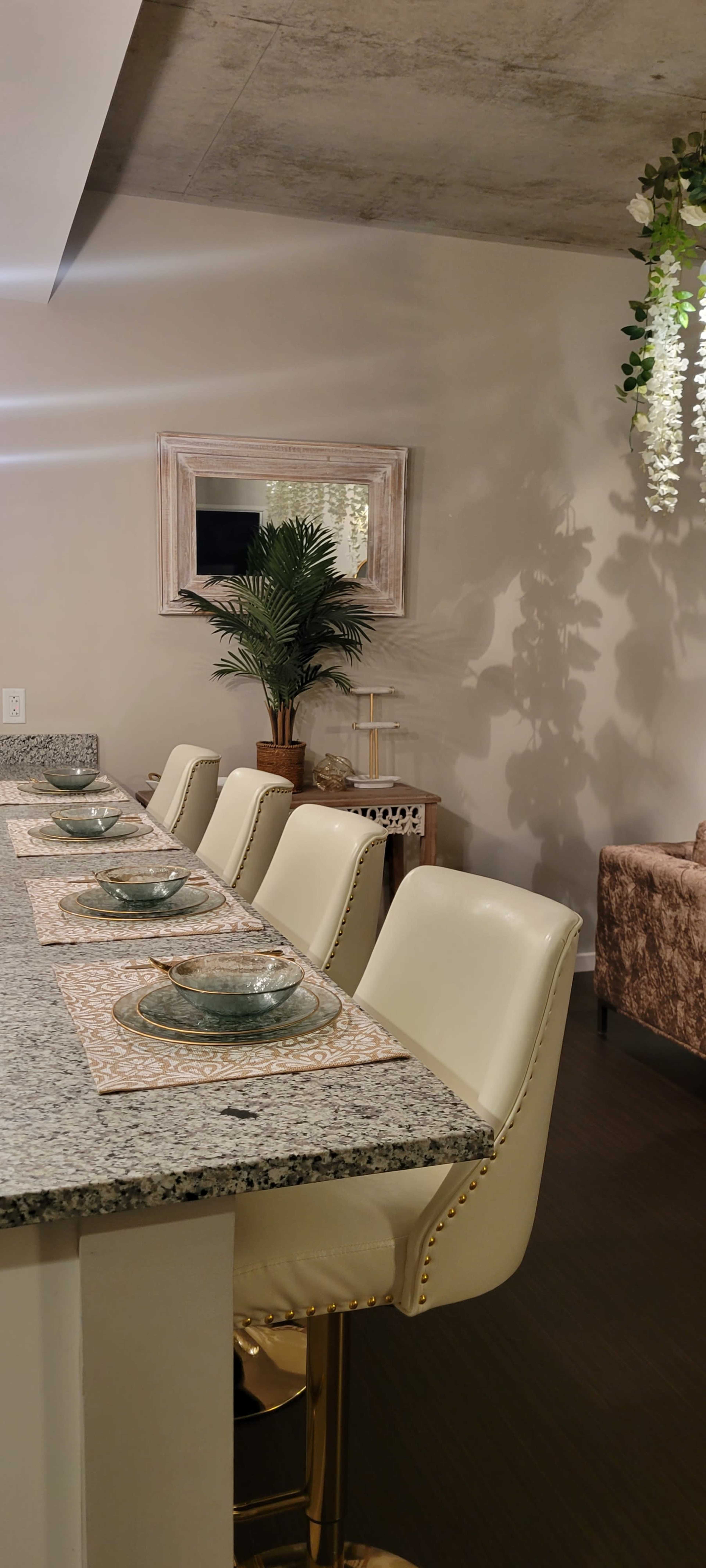 A modern dining area features a granite countertop with six white upholstered stools, elegant table settings, and a decorative plant in the background.