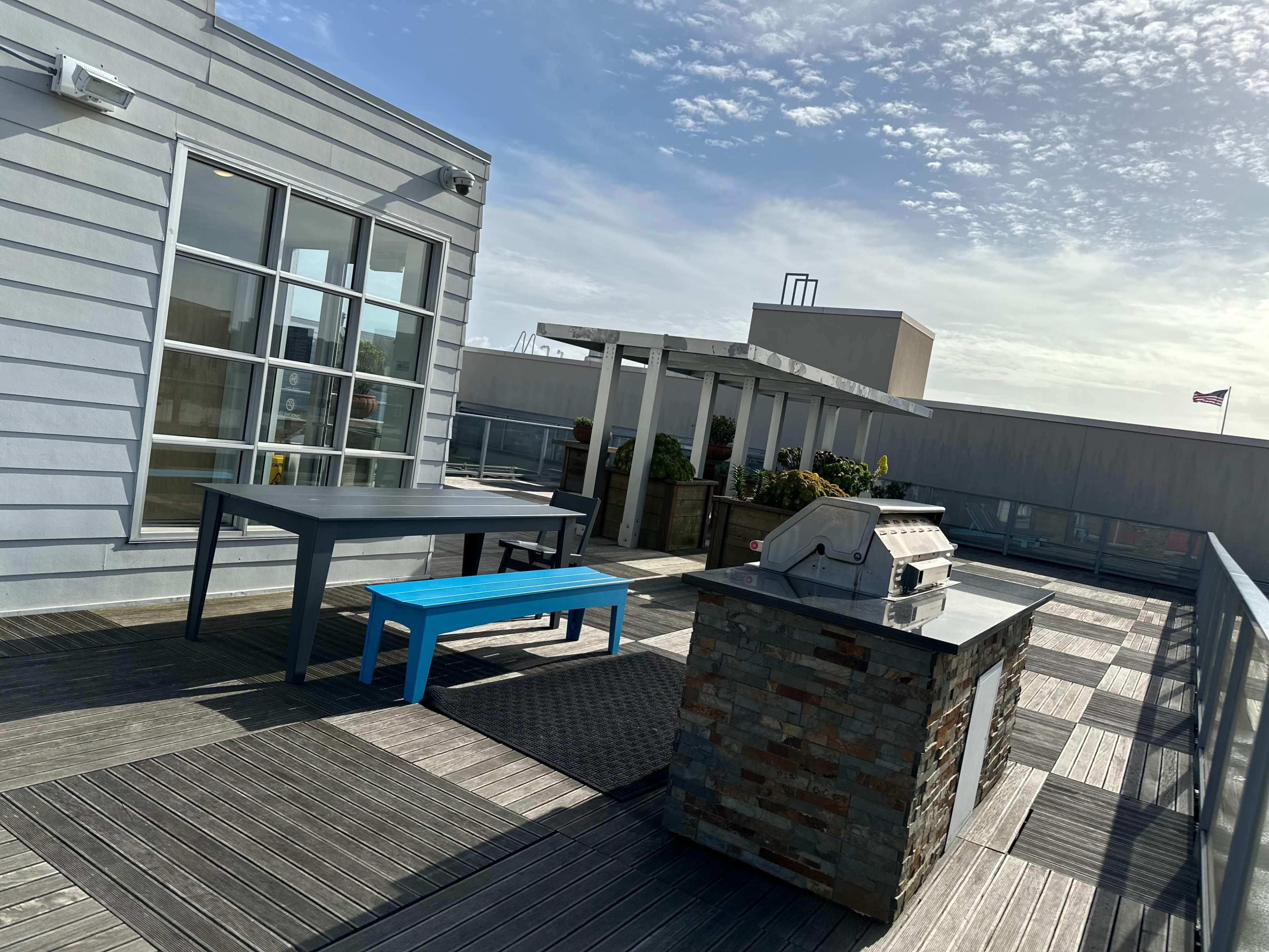 A rooftop terrace featuring a barbecue grill, a stone island, a blue table with benches, and planters surrounded by a wooden deck.
