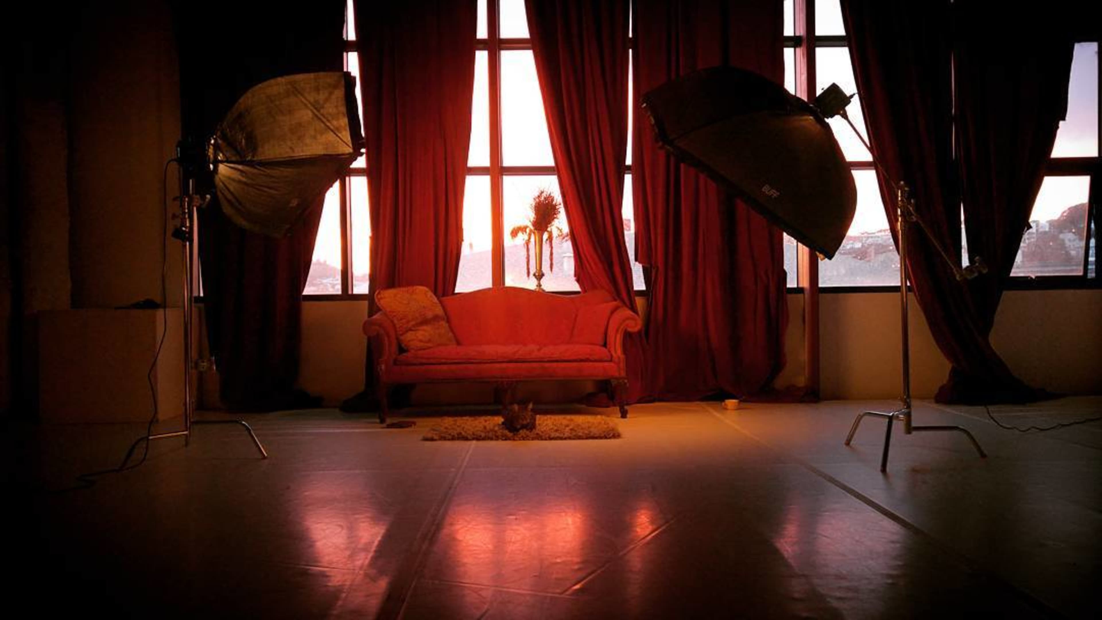 A pink couch is positioned in front of large windows, framed by red curtains, with two studio lights on either side.