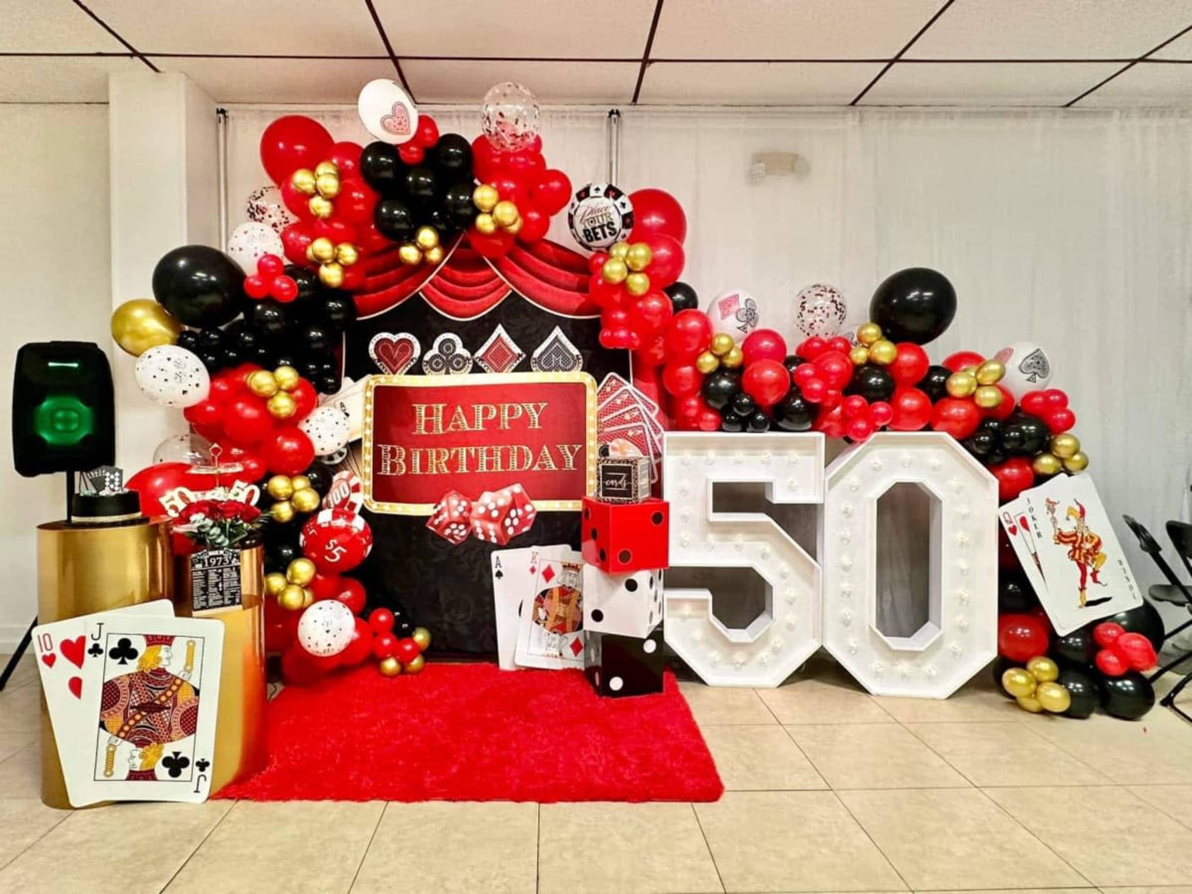 The image features a birthday celebration backdrop decorated with red, black, and gold balloons, a large "50" sign, and playing card motifs.