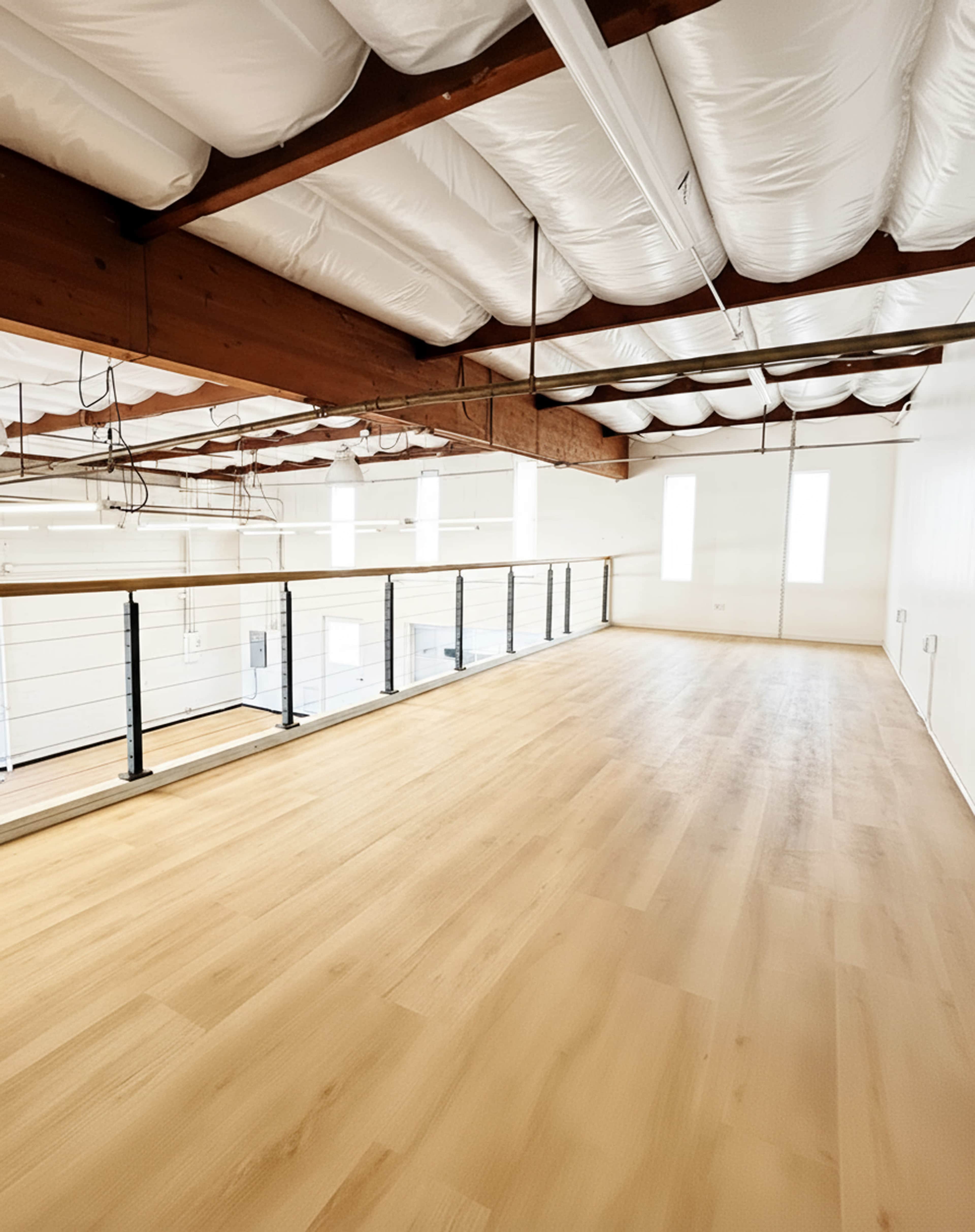 The image shows a spacious, empty room with wooden flooring, large windows, and exposed beams on the ceiling.
