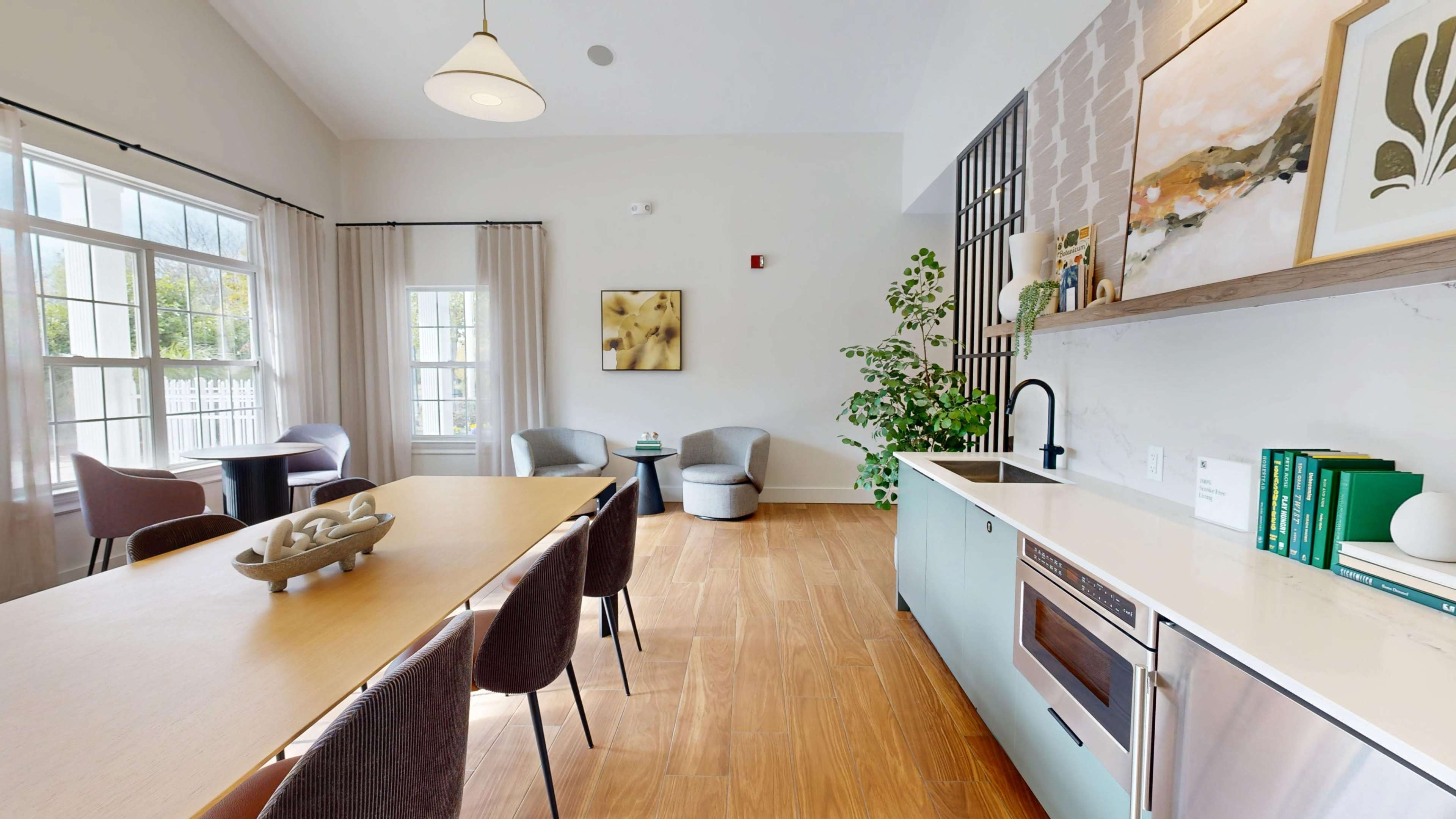 The image shows a modern interior space featuring a dining area with a long table, a kitchenette, and two armchairs beside a window.