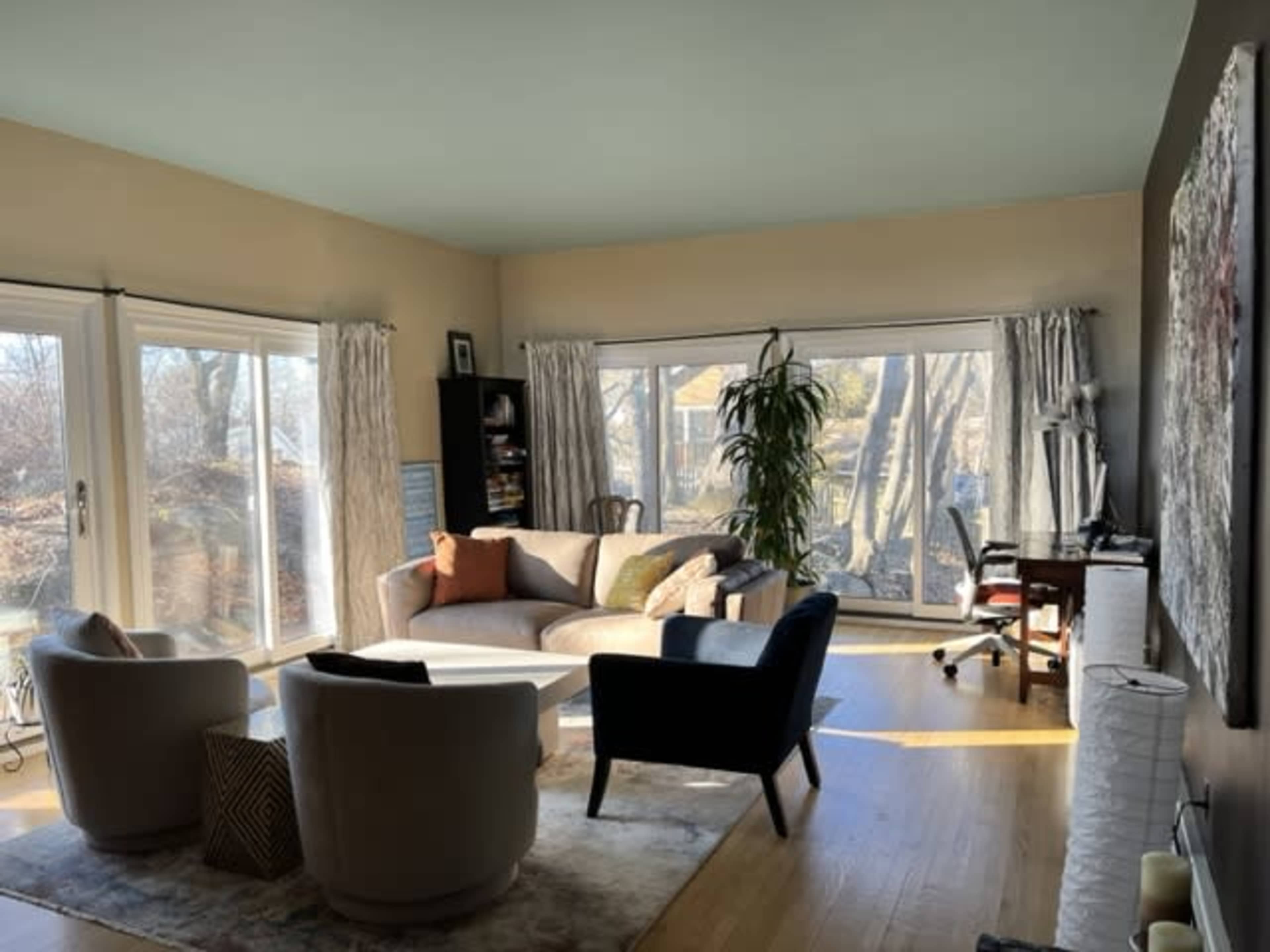 A bright living room features large windows, a curved sofa, two armchairs, and a desk near the corner.