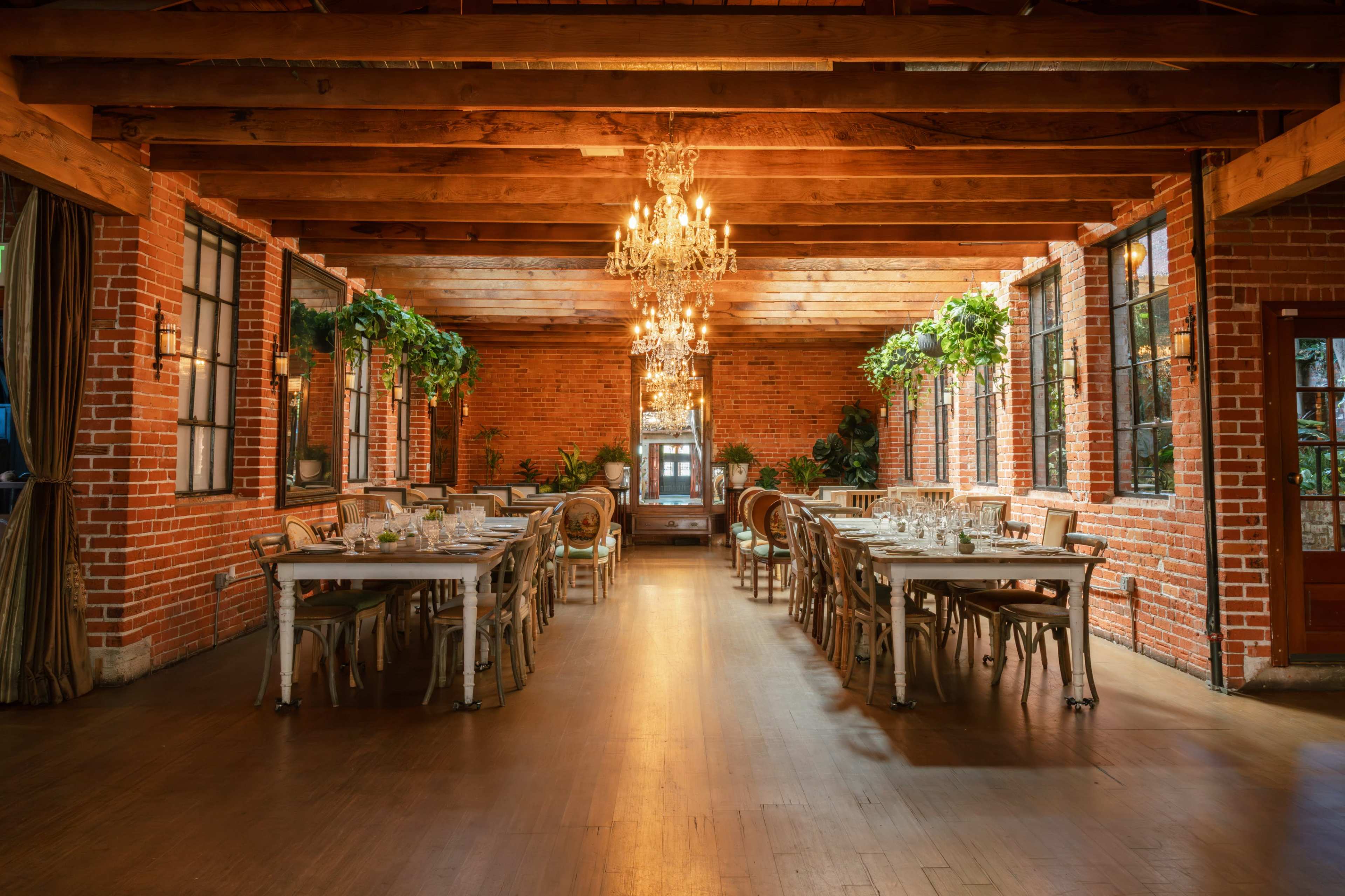 The image depicts a spacious dining area with wooden beams, exposed brick walls, and elegant chandeliers, featuring tables set for dining and surrounded by plants.