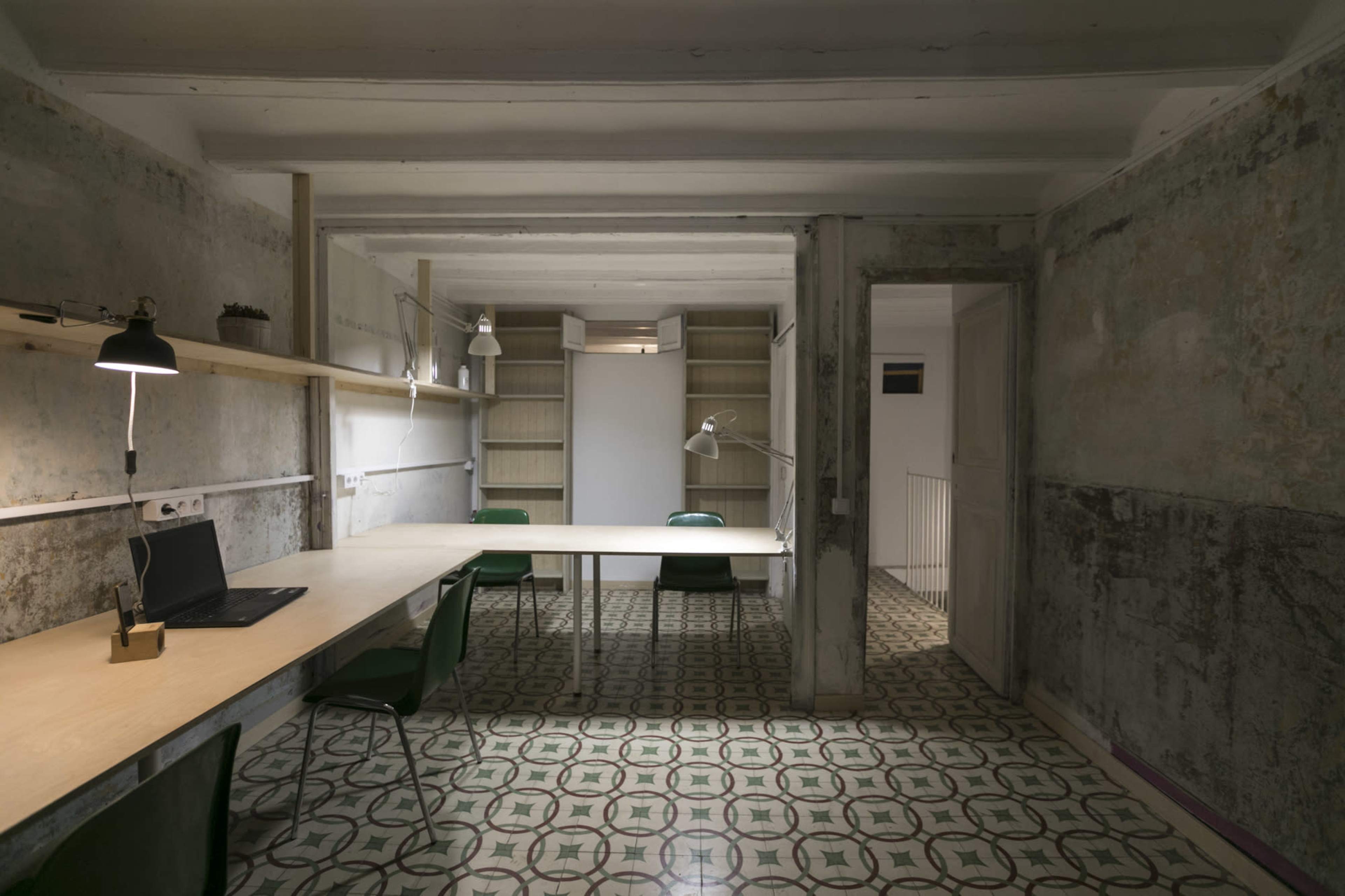 The image shows a sparsely furnished room with a long wooden table, green chairs, and shelves against weathered walls, featuring tiled flooring.