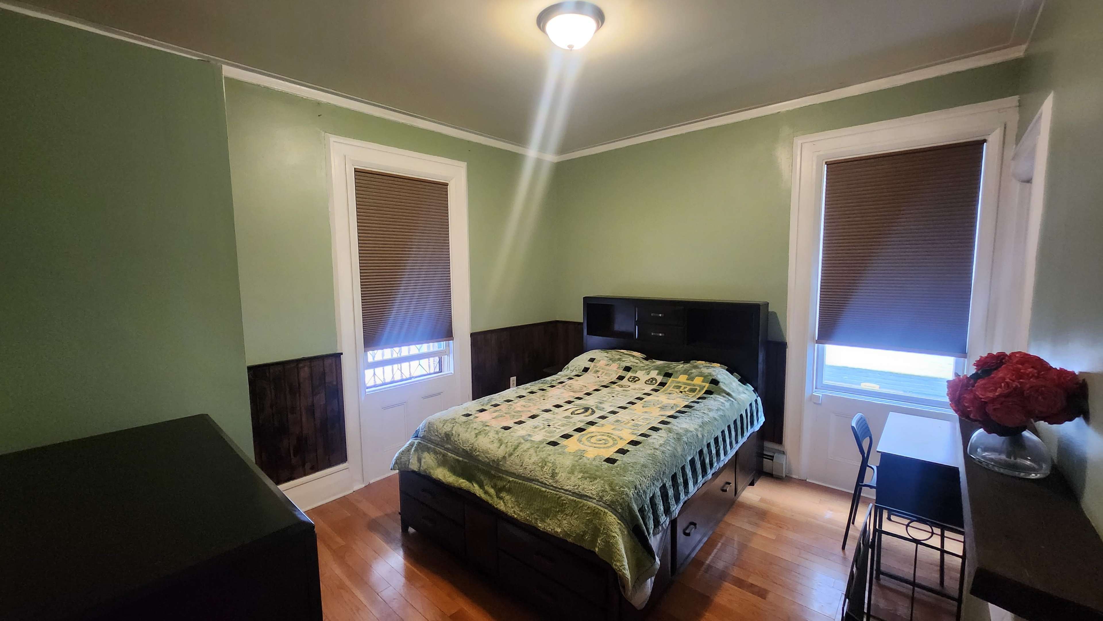 A small bedroom with green walls, a bed covered with a patterned blanket, a desk with a chair, and two windows with brown shades.