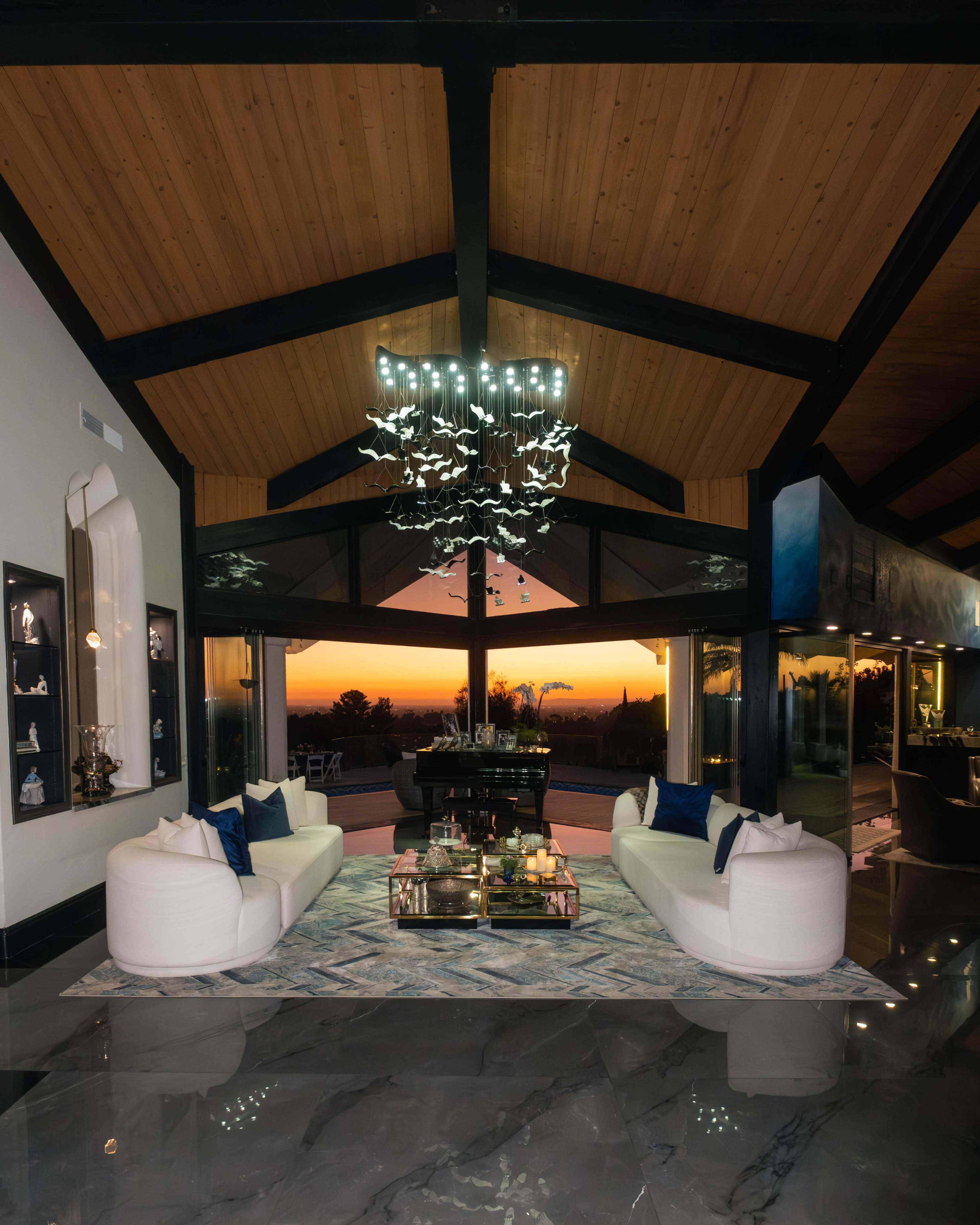 The image shows a modern living room with two white sofas facing a glass coffee table, large windows revealing a sunset view, and a chandelier made of abstract shapes.