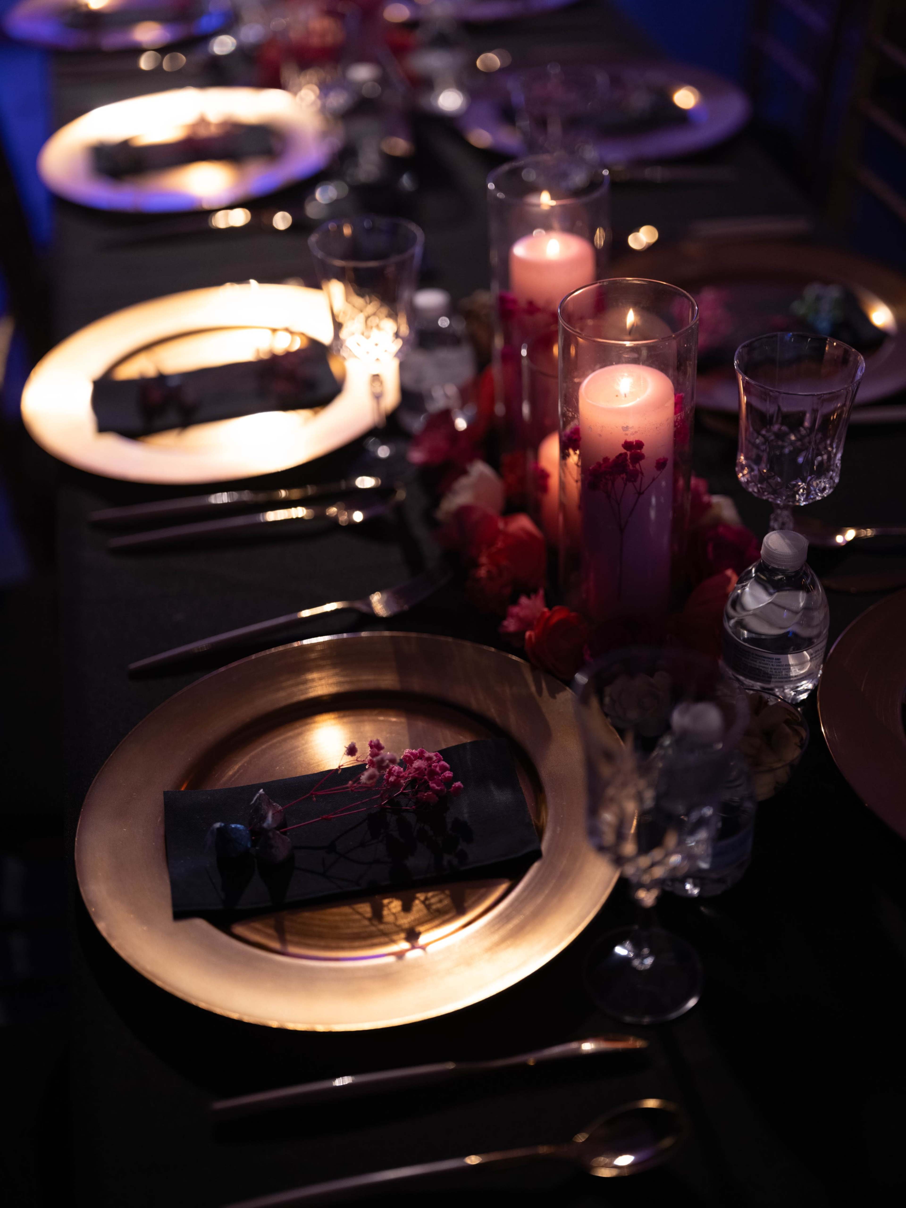 A formal dining table is set with gold plates, black napkins, and glowing candles surrounded by floral arrangements.