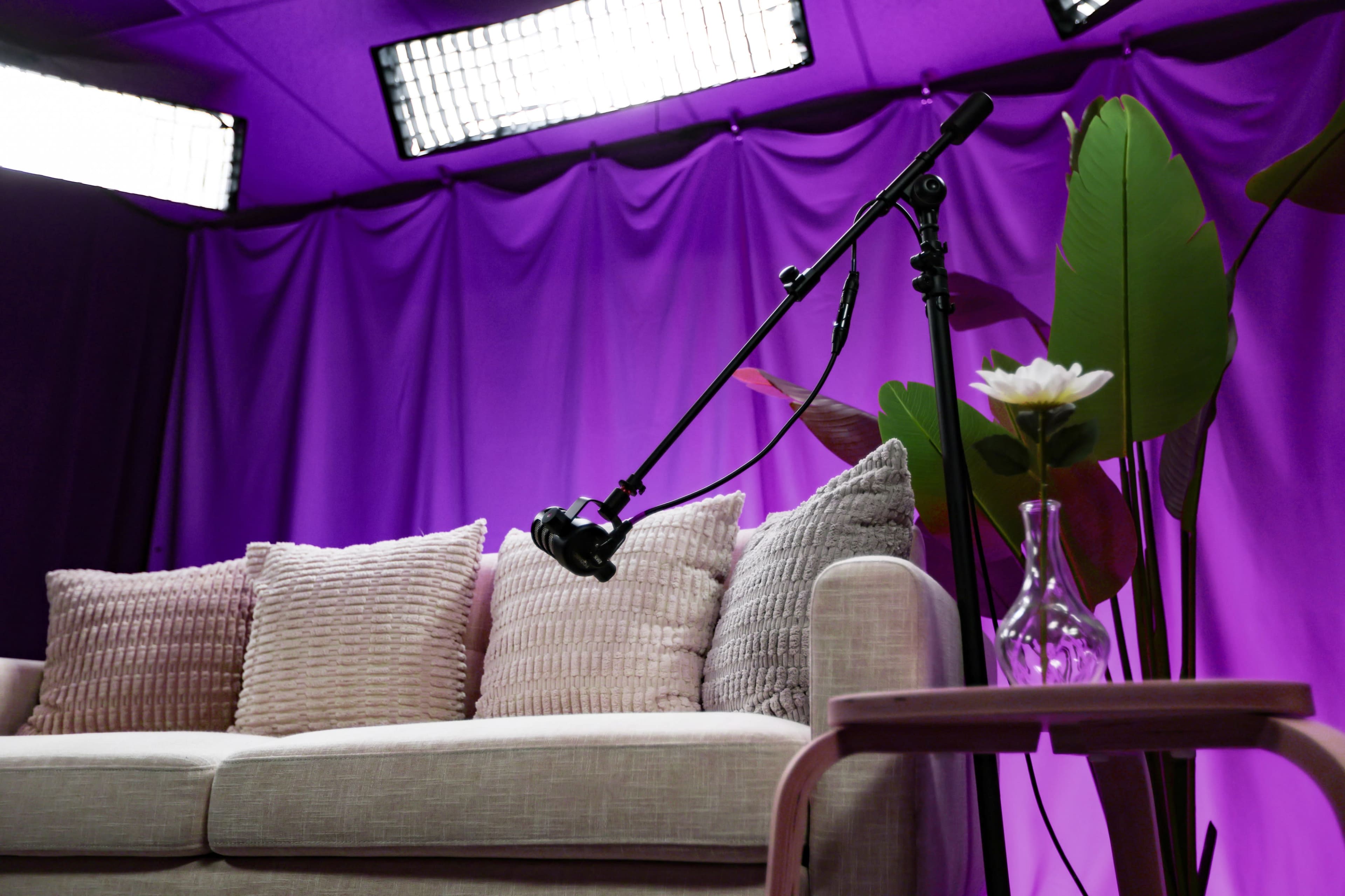 A microphone is positioned on a stand beside a light-colored sofa adorned with decorative pillows, set against a purple backdrop with studio lights.