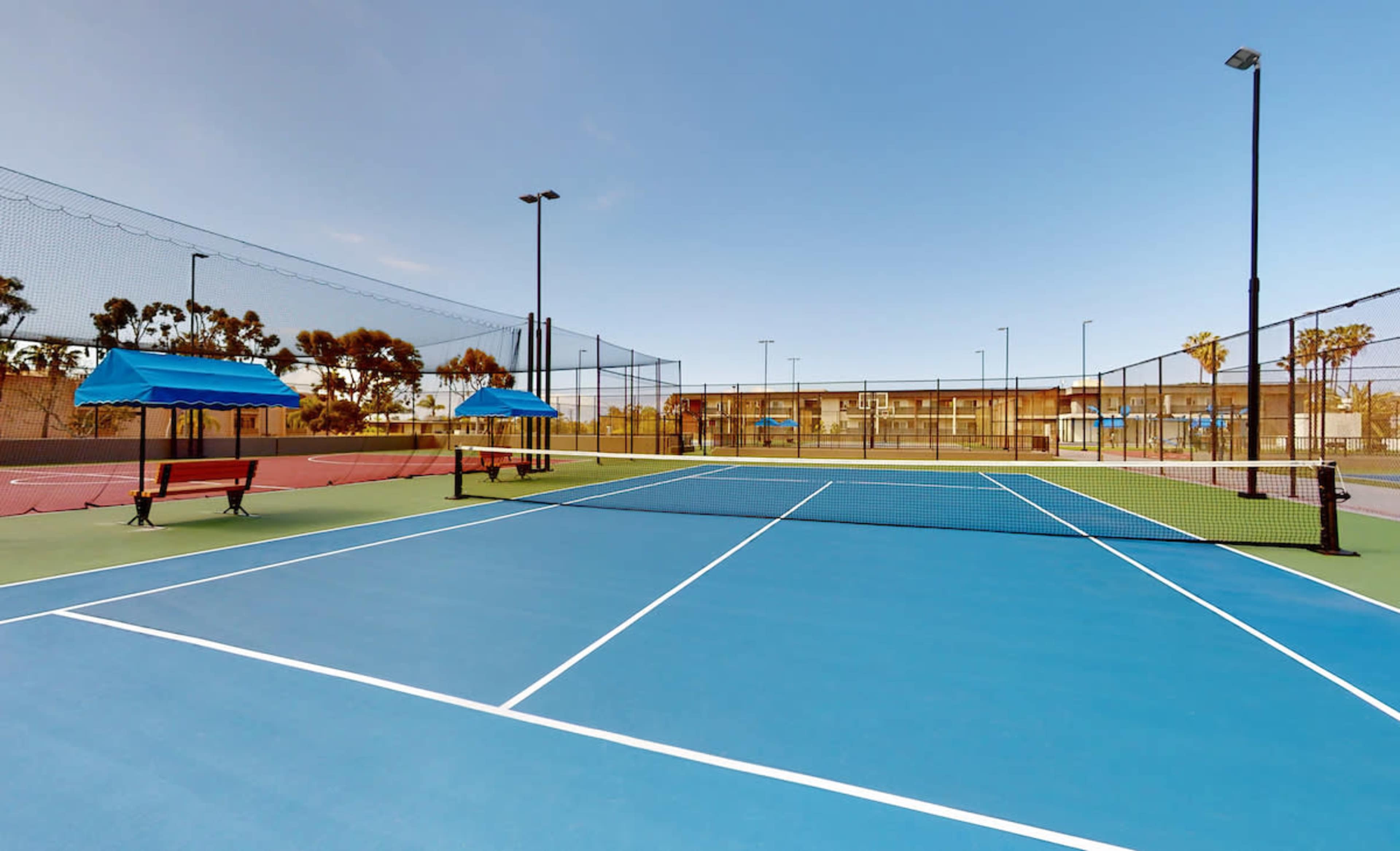 A well-maintained tennis court with two blue canopies and benches, surrounded by additional courts and palm trees in the background.