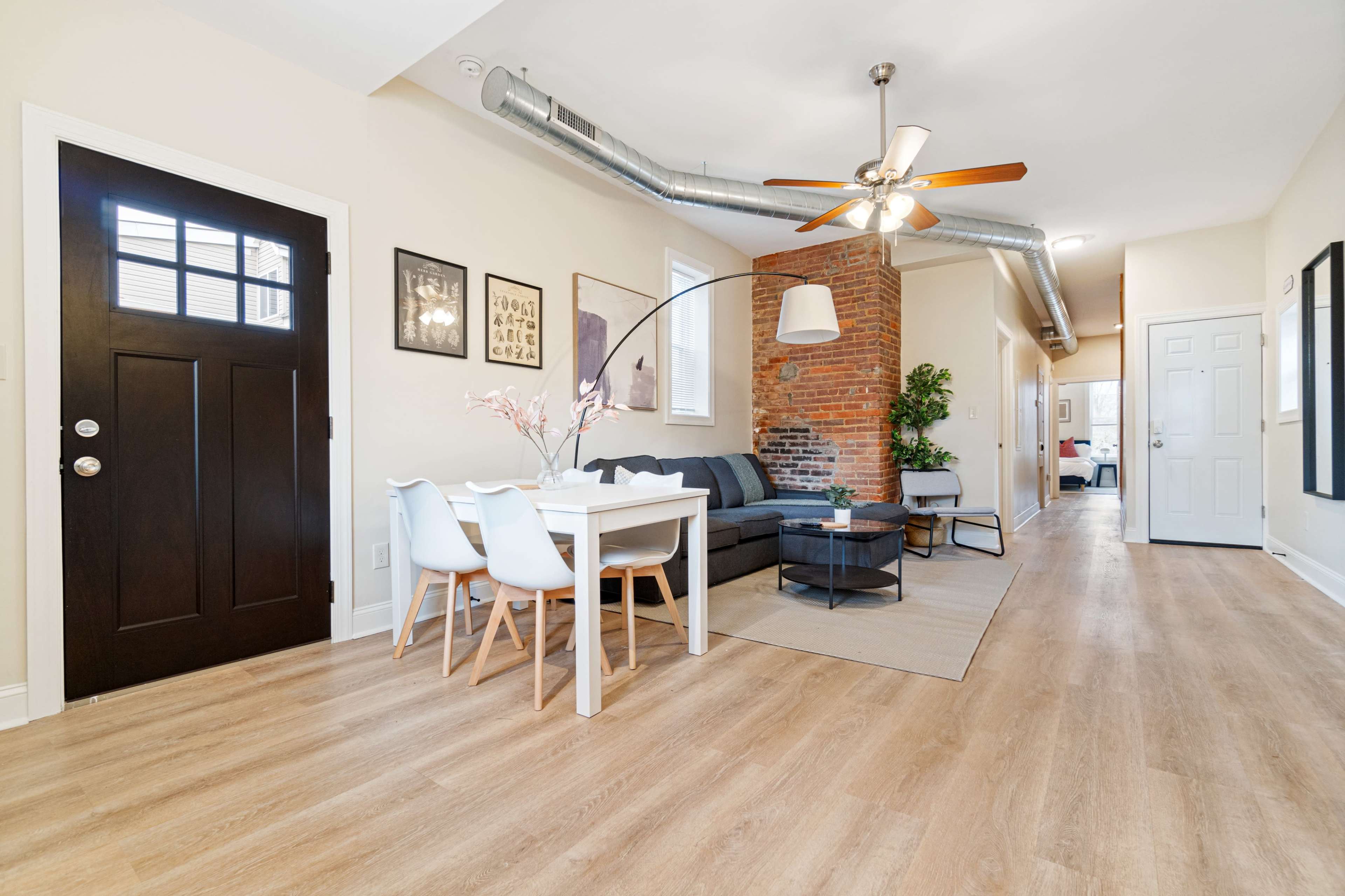 The image shows a modern living space with a dining table and chairs, a sectional sofa, and a brick accent wall, leading to a hallway with doors.