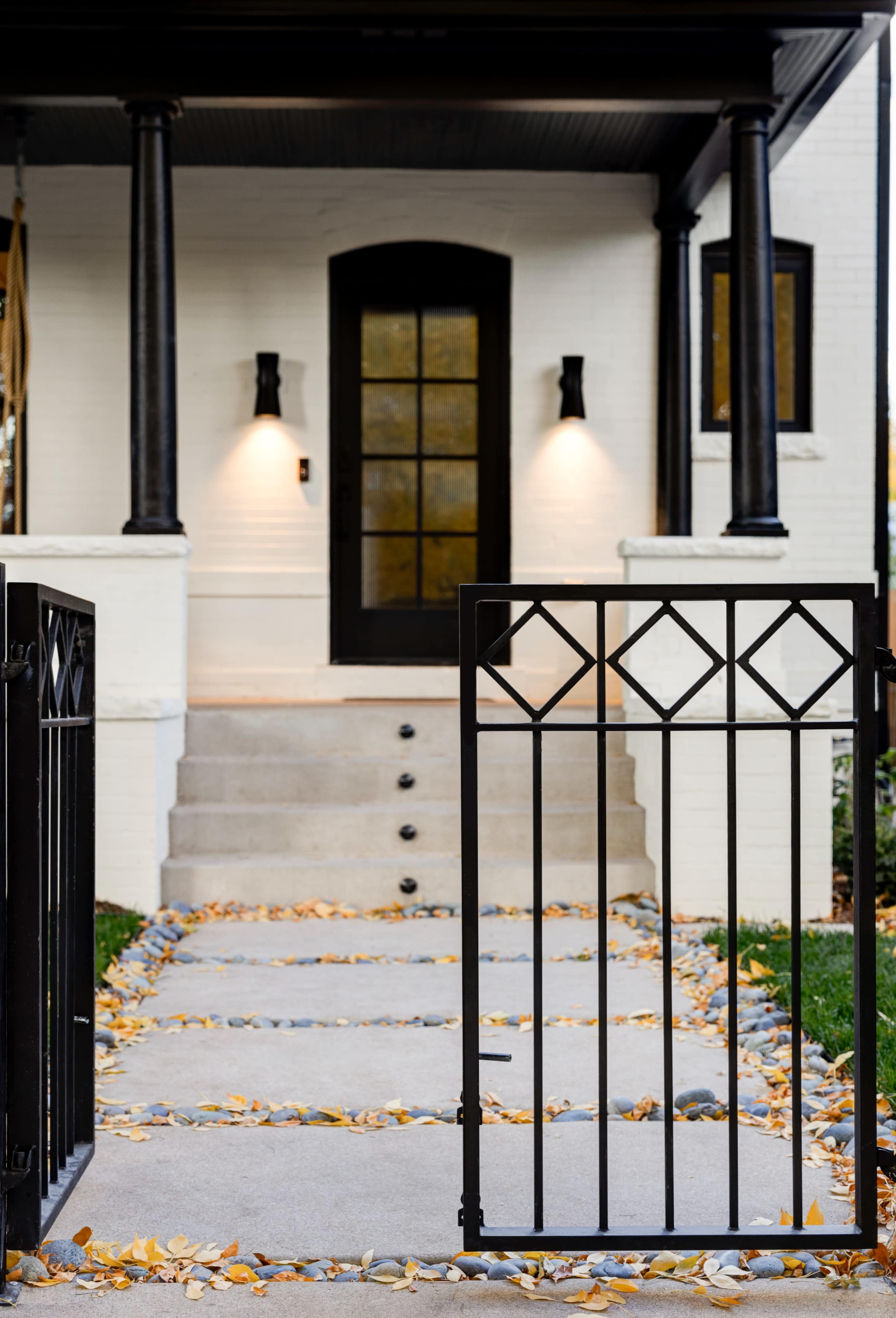 A pathway lined with fallen leaves leads to the entrance of a white house with a dark front door and exterior lighting.