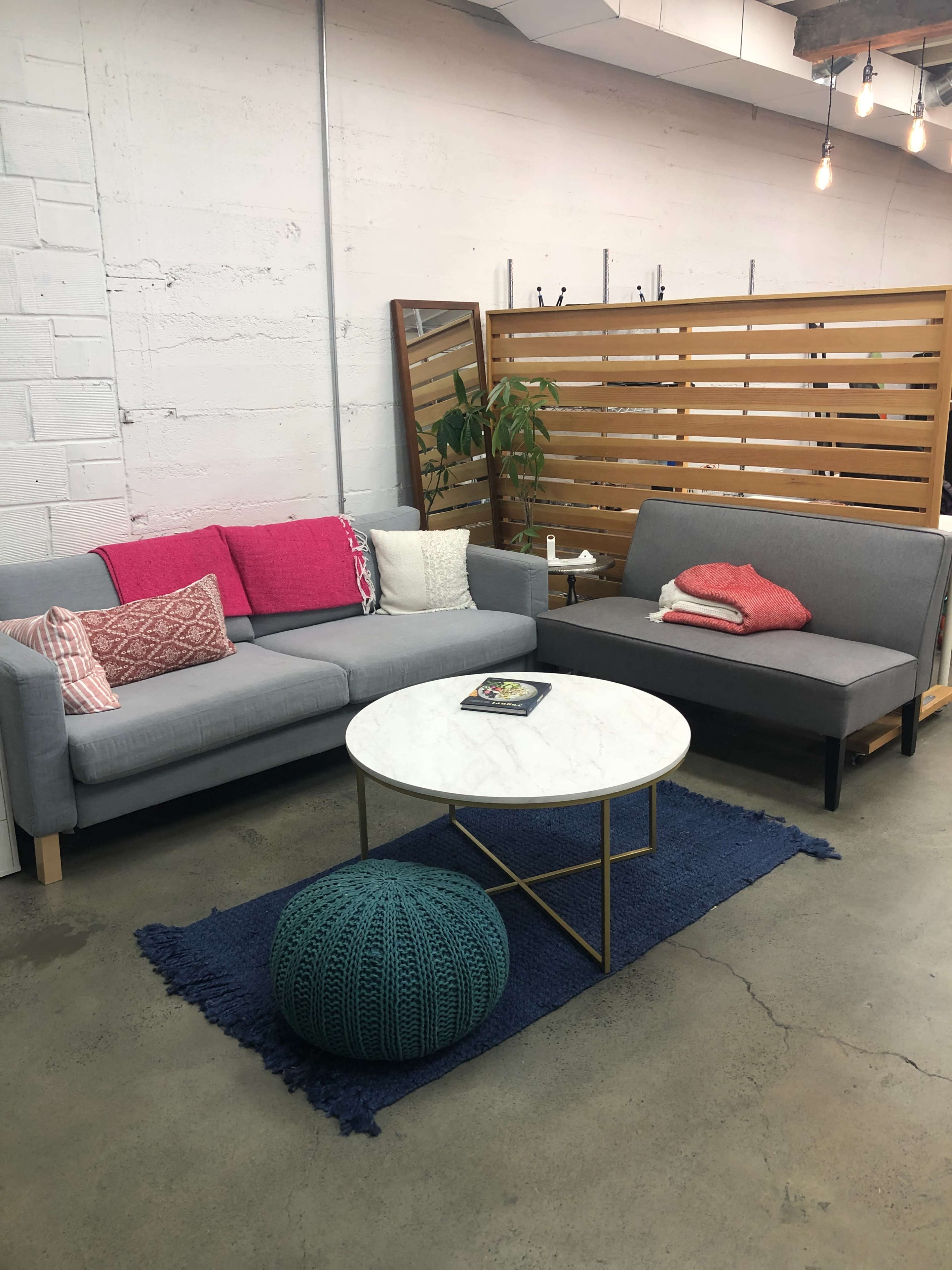A modern living room area featuring a gray sofa and a gray loveseat, a round marble coffee table, and a knitted pouf, all arranged on a blue rug against a textured wall.