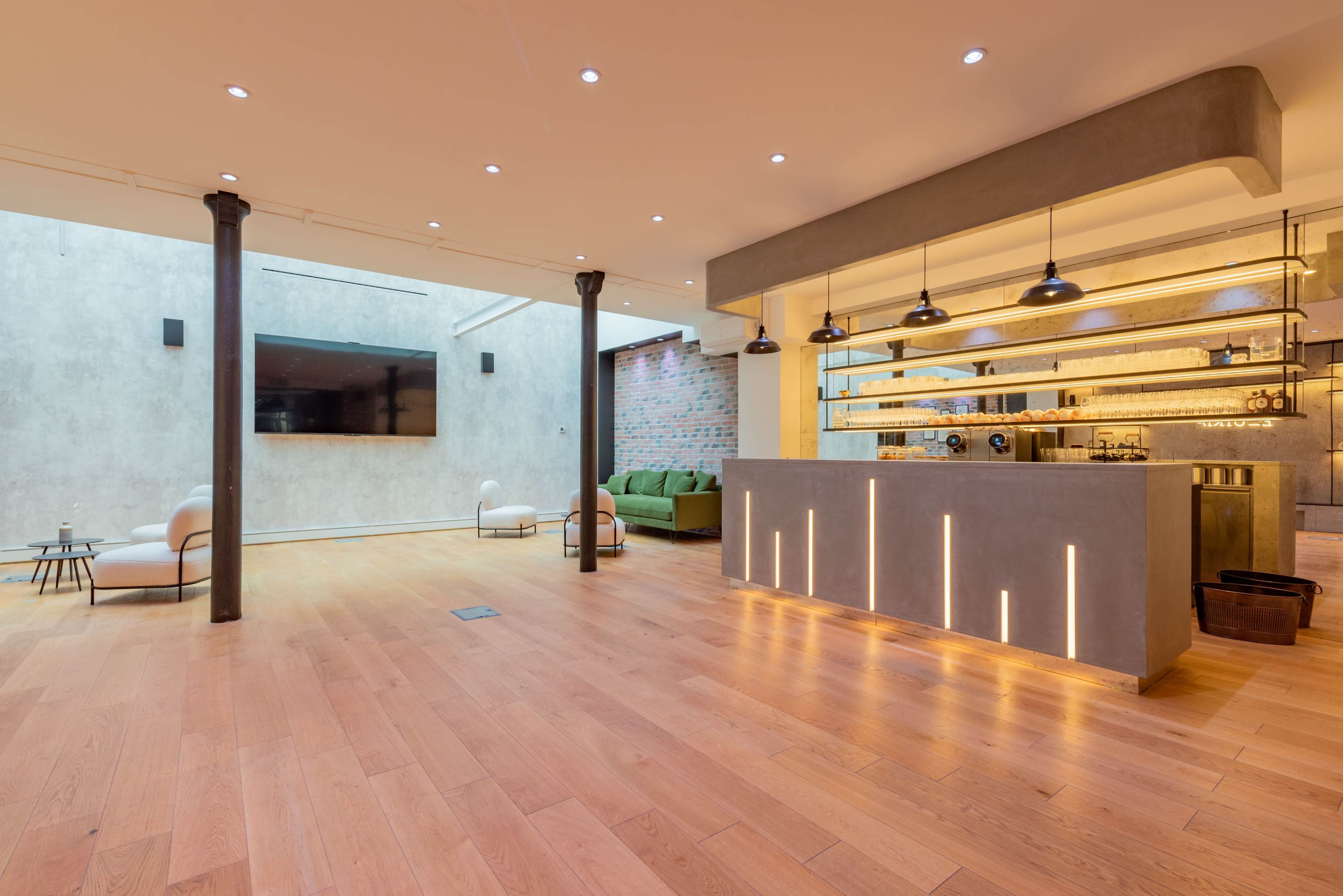 The image shows a spacious, modern interior with wooden flooring, a minimalist bar area, and a green seating arrangement against a textured wall.