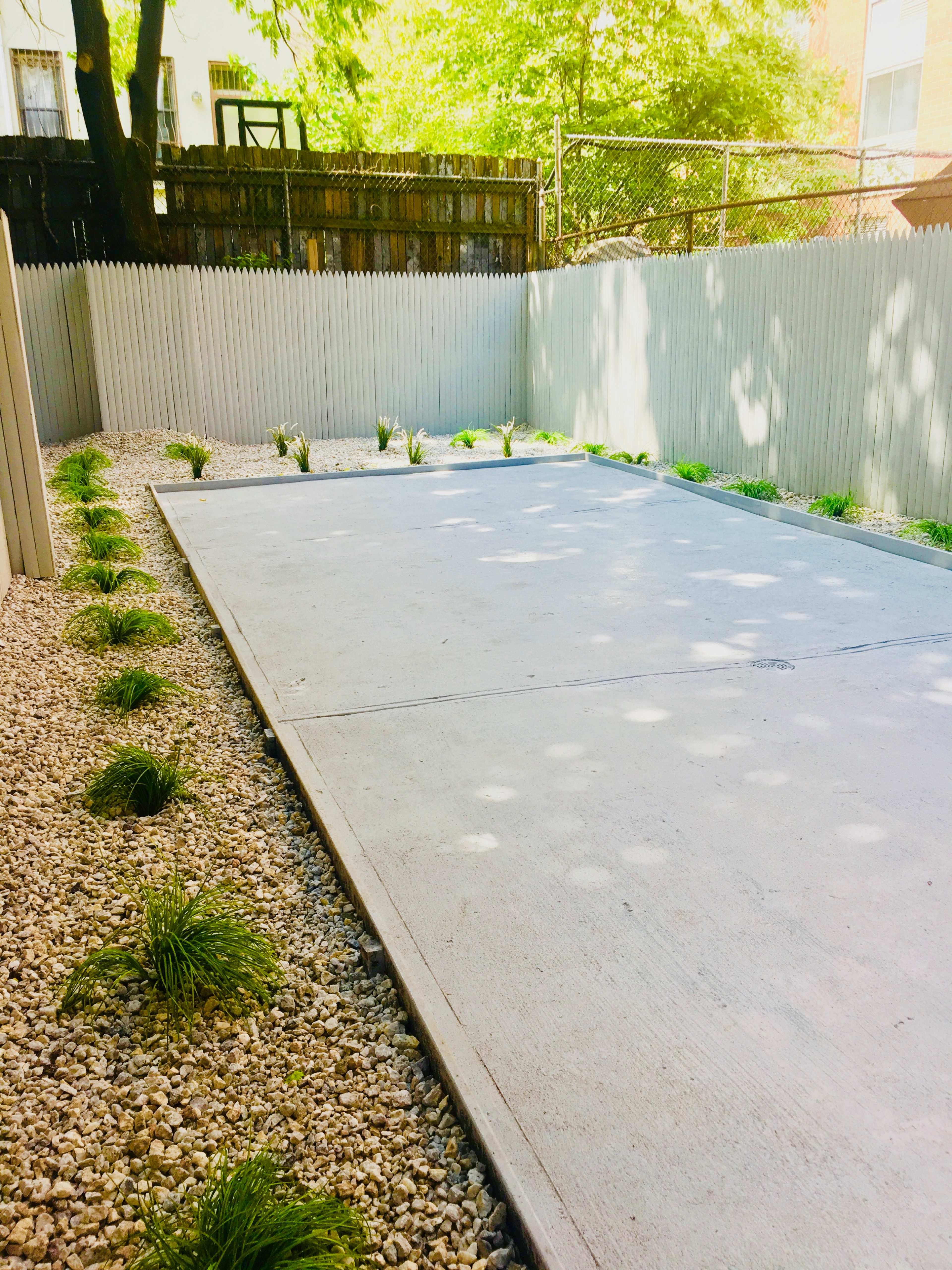 A paved area surrounded by gravel and small plants, bordered by a white wooden fence in a sunny outdoor space.