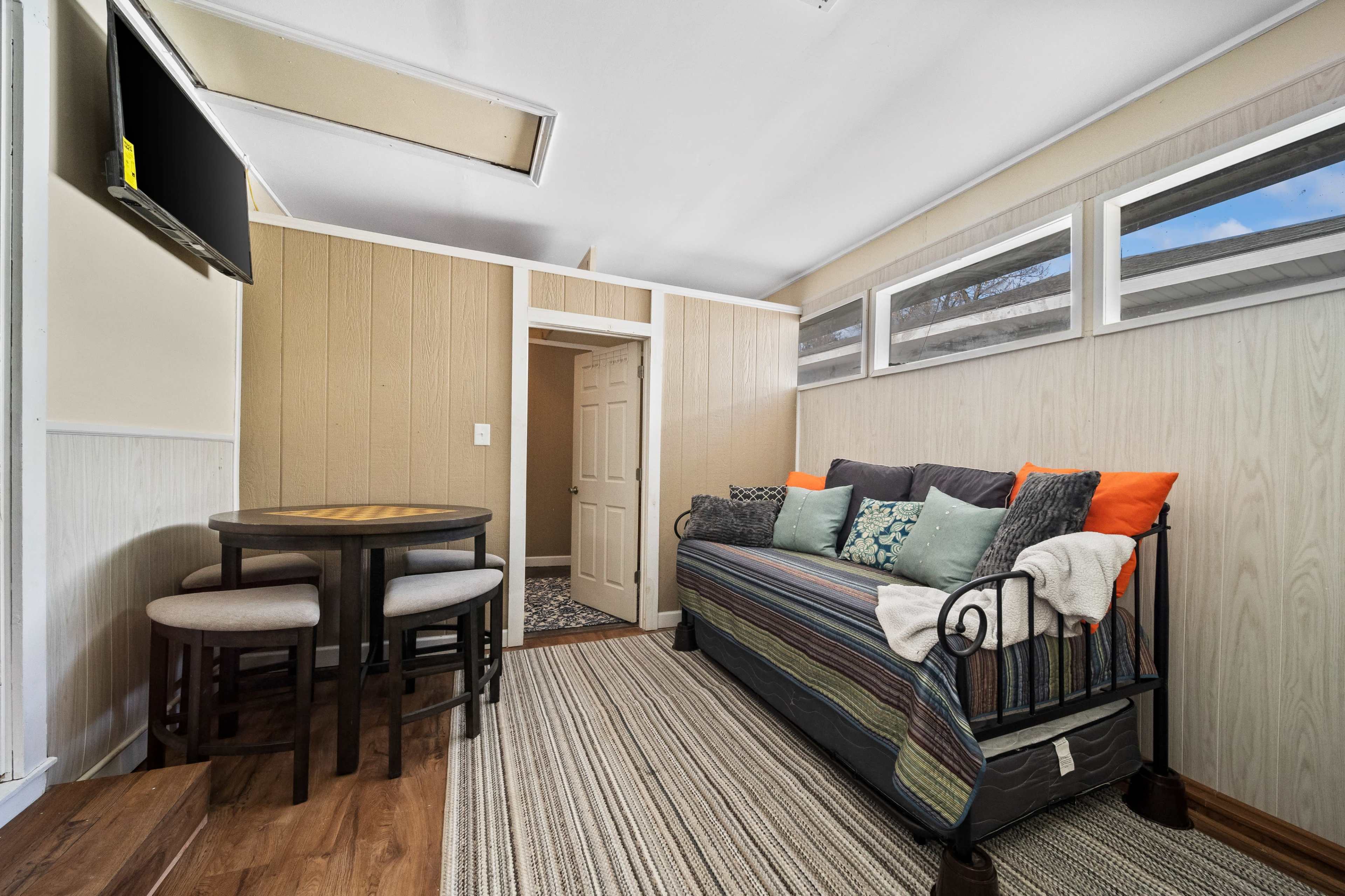 The image shows a simple room with a small dining table and chairs, a couch with colorful cushions, and a television mounted on the wall.