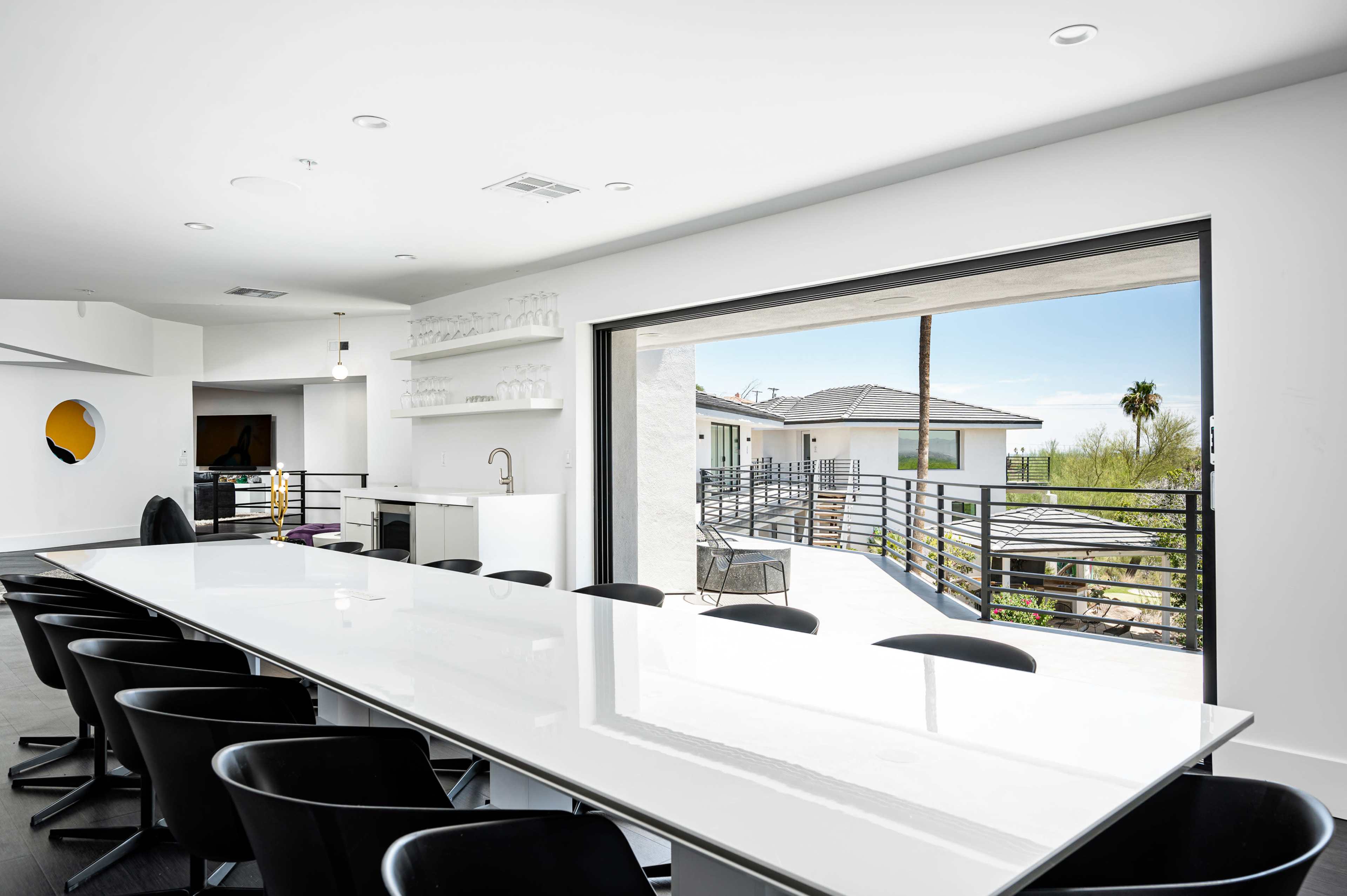 A modern dining area features a long white table and large sliding doors that open to a balcony with a view of palm trees and a clear sky.
