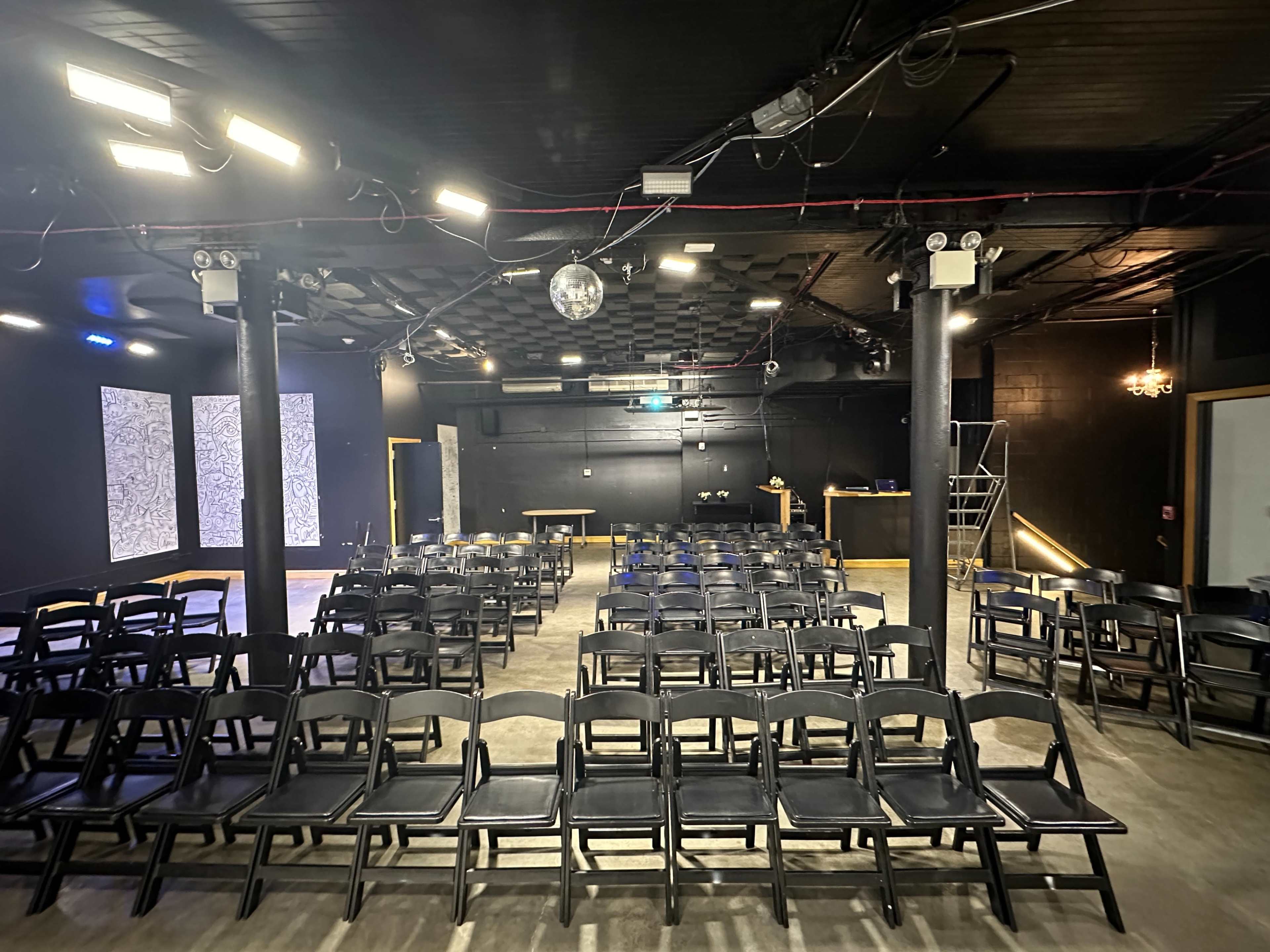 The image shows a sparsely populated theater space with rows of black folding chairs facing a small stage illuminated by stage lights.