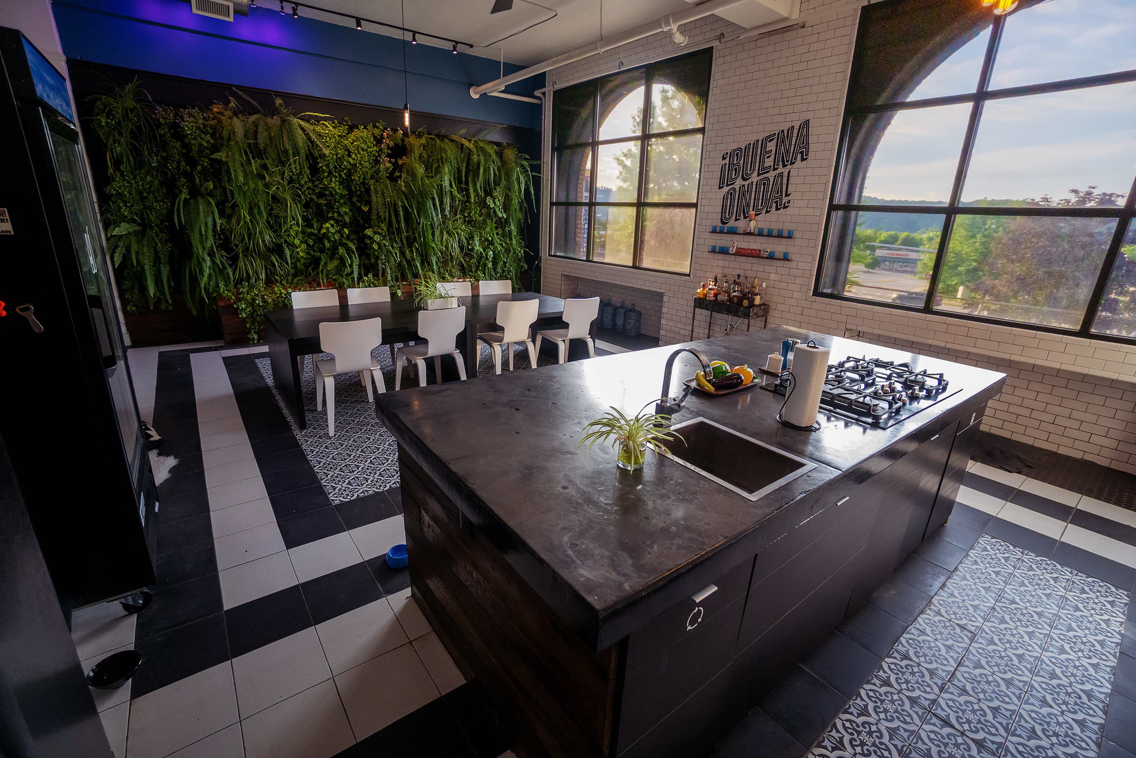 A modern kitchen features a large island with a sink and stove, surrounded by white chairs and a wall of greenery, alongside large windows that provide natural light.