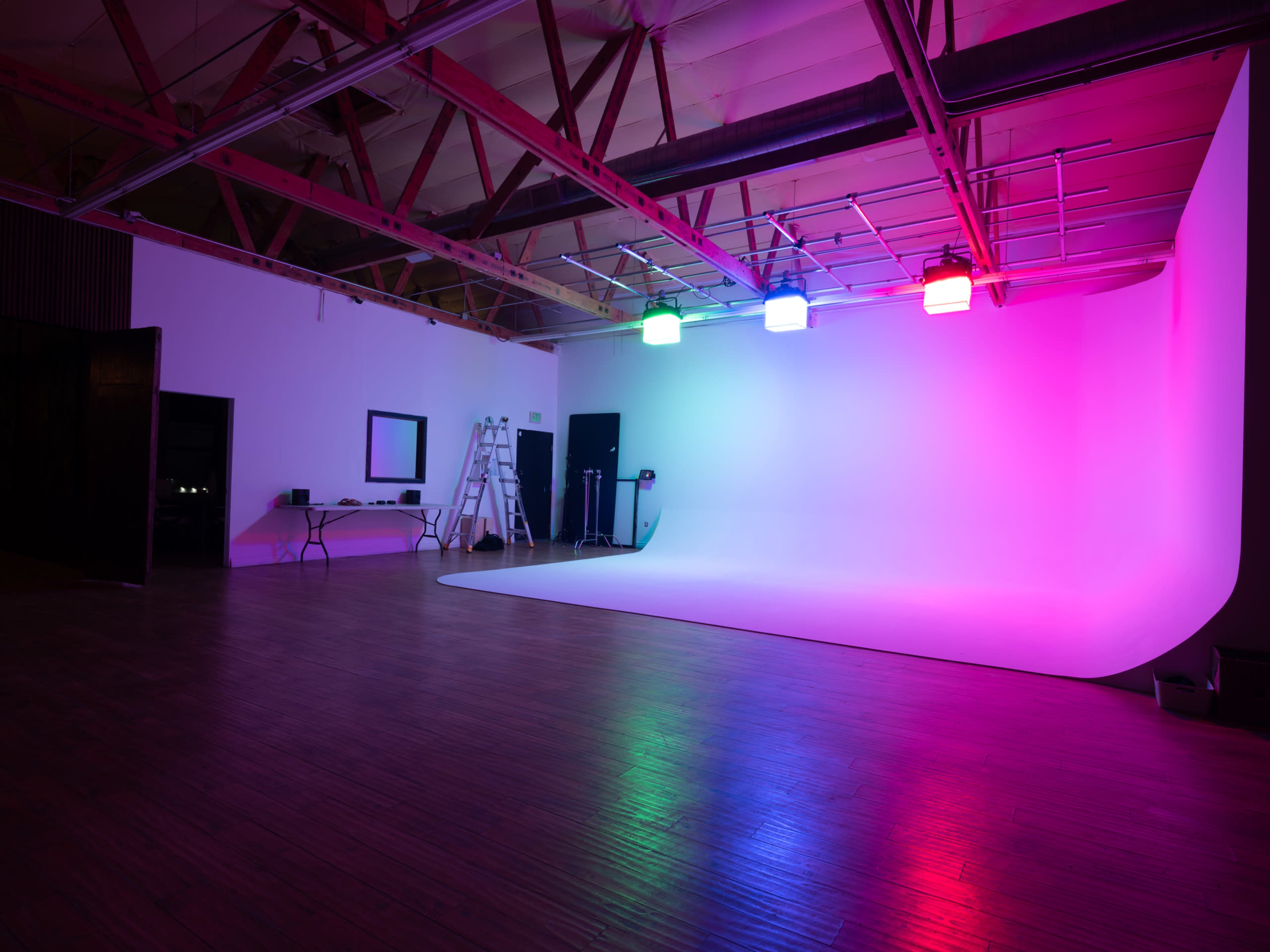 A spacious photography studio features a large white backdrop with colored lighting in pink, green, and blue, and a ladder and equipment set against the walls.