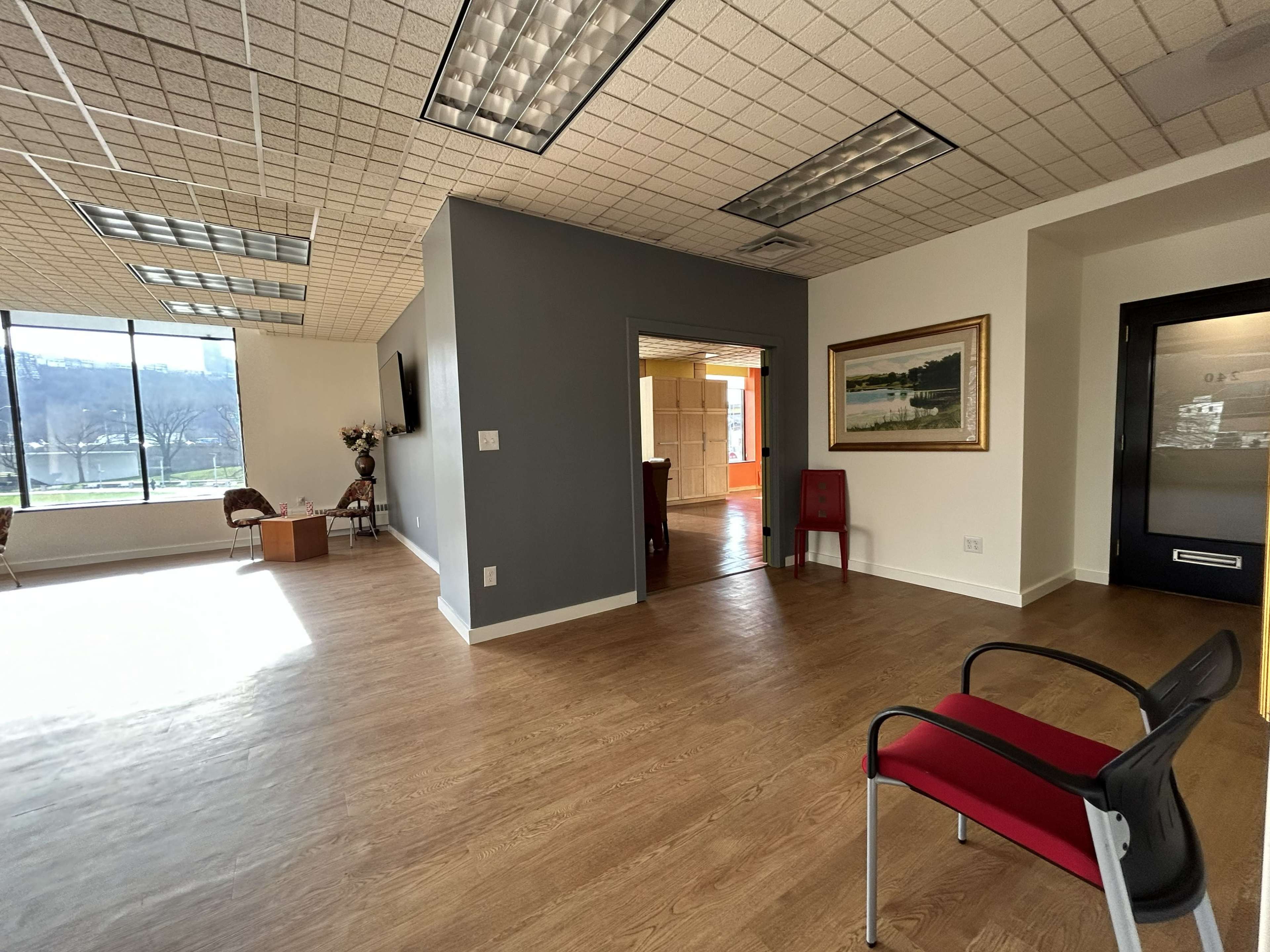 The image depicts a spacious office interior with wooden flooring, a waiting area featuring two chairs, and a large window allowing natural light to enter.