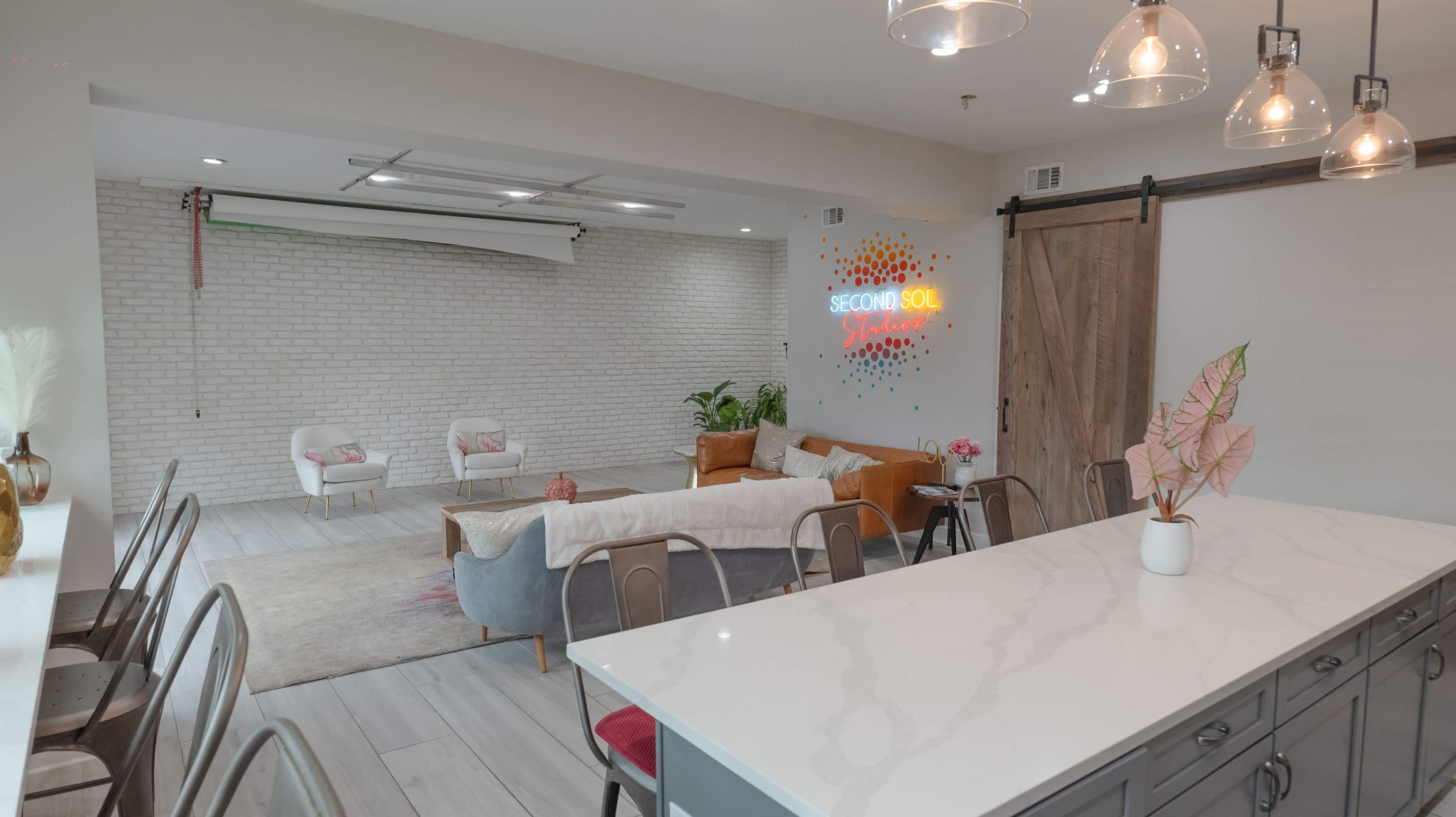 A modern, open-concept living space featuring a seating area with a gray couch, wooden chairs, and a neon sign on the wall, with a kitchen island visible in the foreground.
