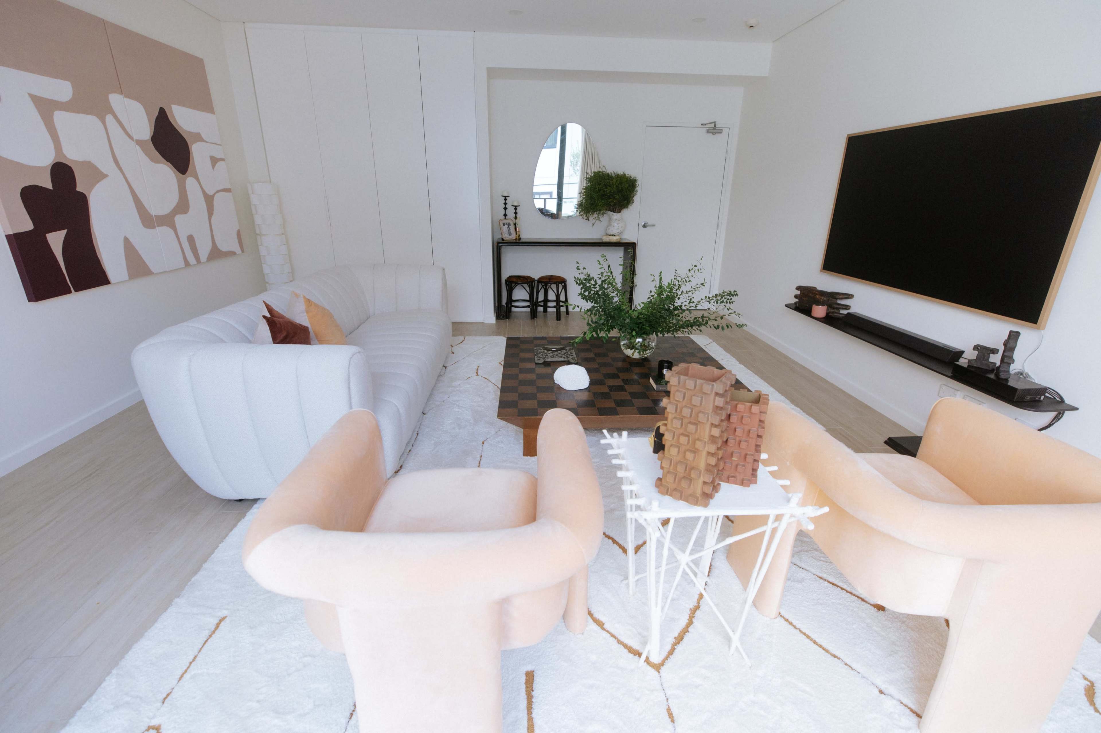 A modern, minimalistic living room featuring a white sofa, two peach-colored armchairs, a coffee table with a checkerboard pattern, and decorative elements on a side table.