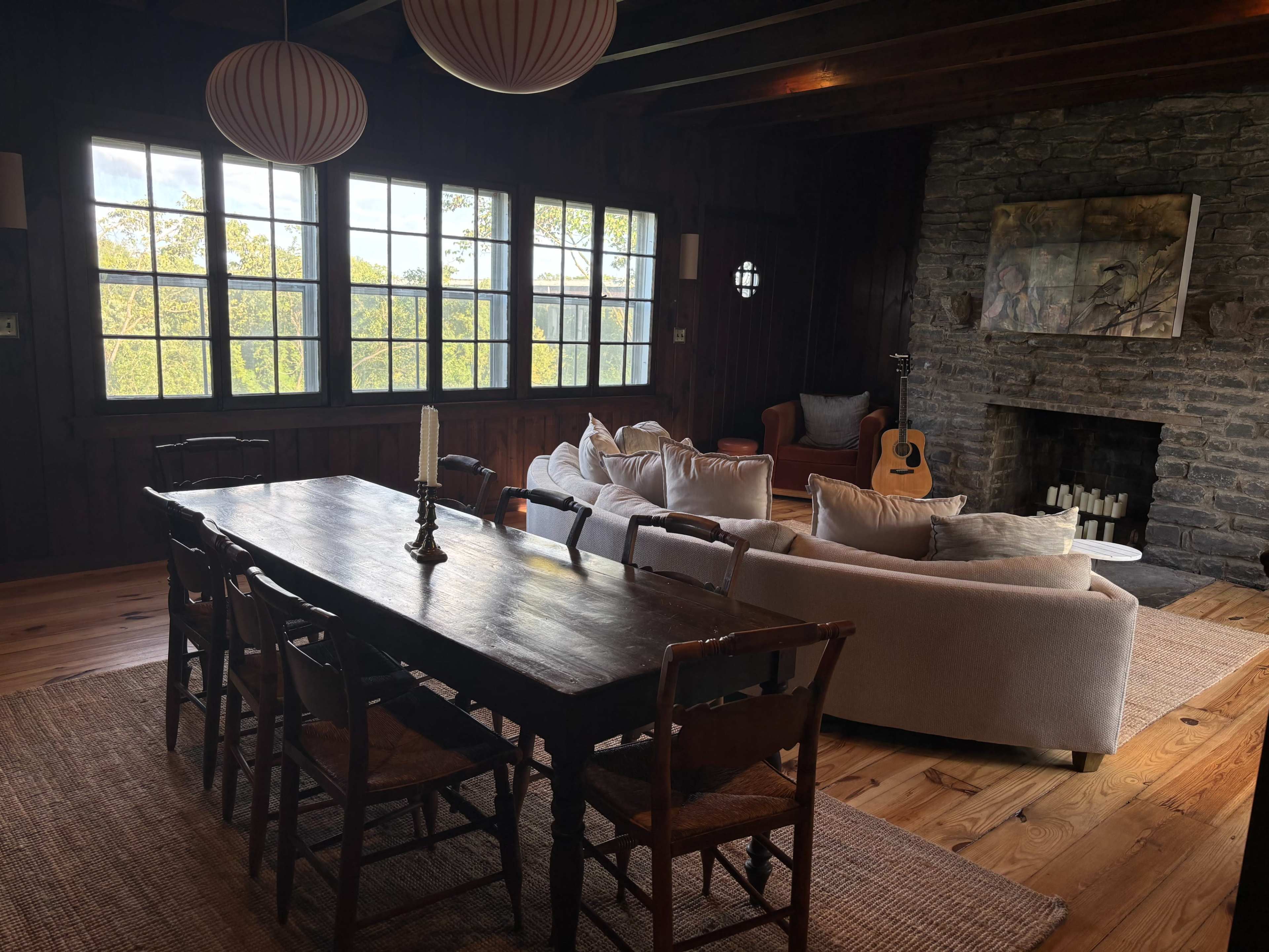 The image shows a rustic interior living space featuring a wooden dining table, a sectional sofa, and large windows revealing a view of greenery outside.