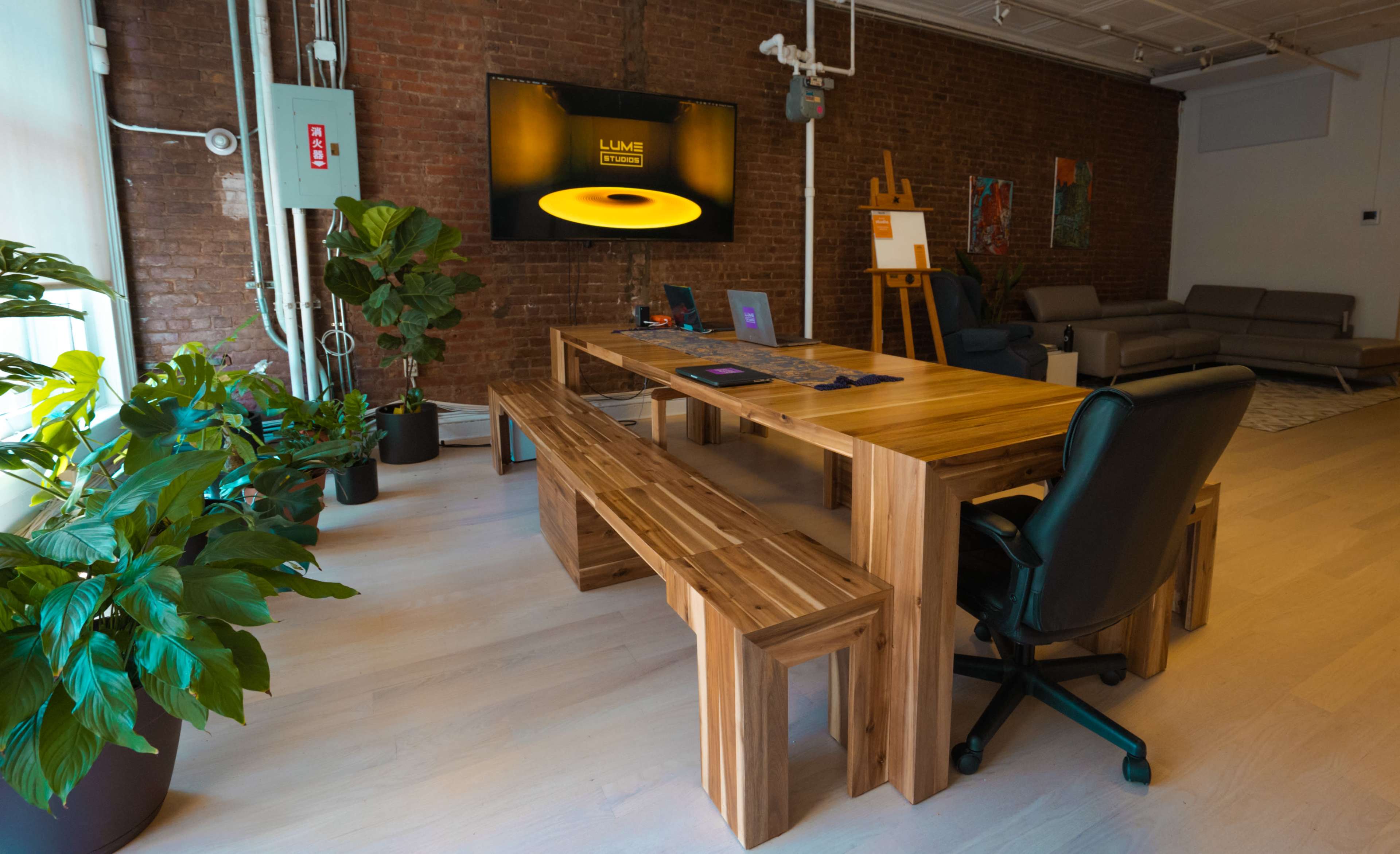 A modern workspace features a large wooden table surrounded by benches and a black office chair, with a TV mounted on the wall displaying a circular logo, and indoor plants in the corners.