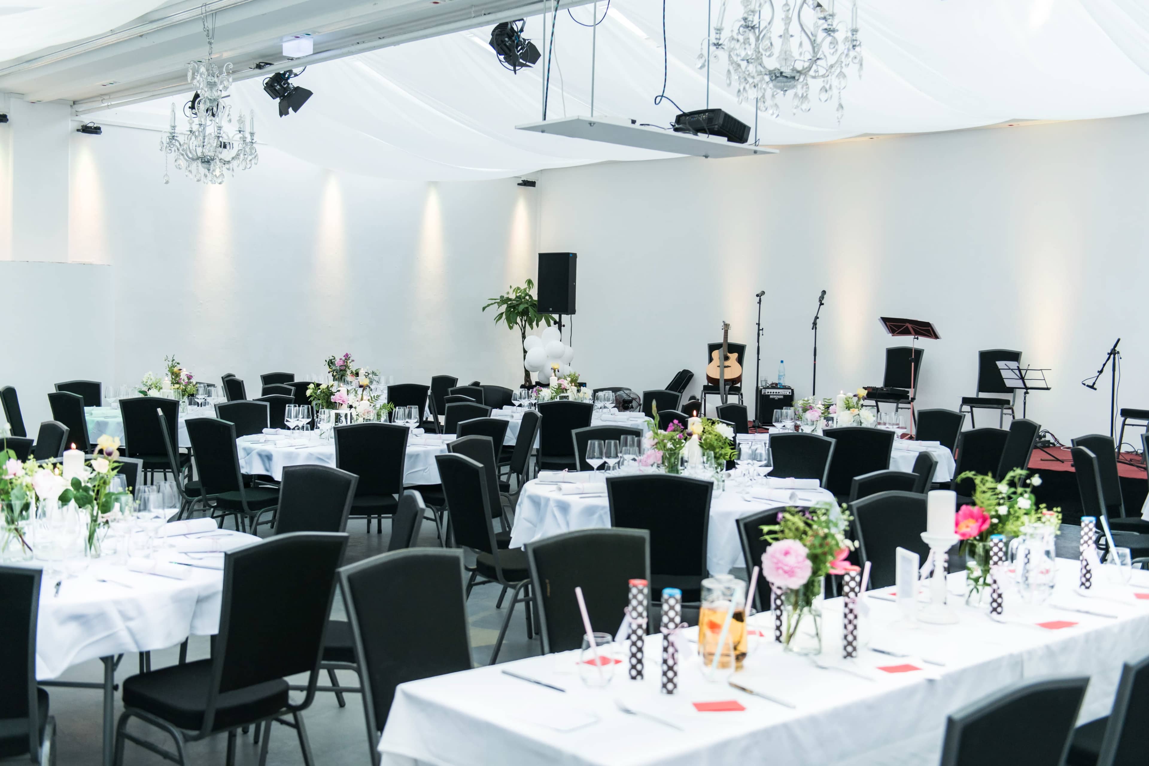 A banquet hall is set with elegantly arranged tables, each adorned with floral centerpieces and surrounded by black chairs, under soft lighting and decorative chandeliers.