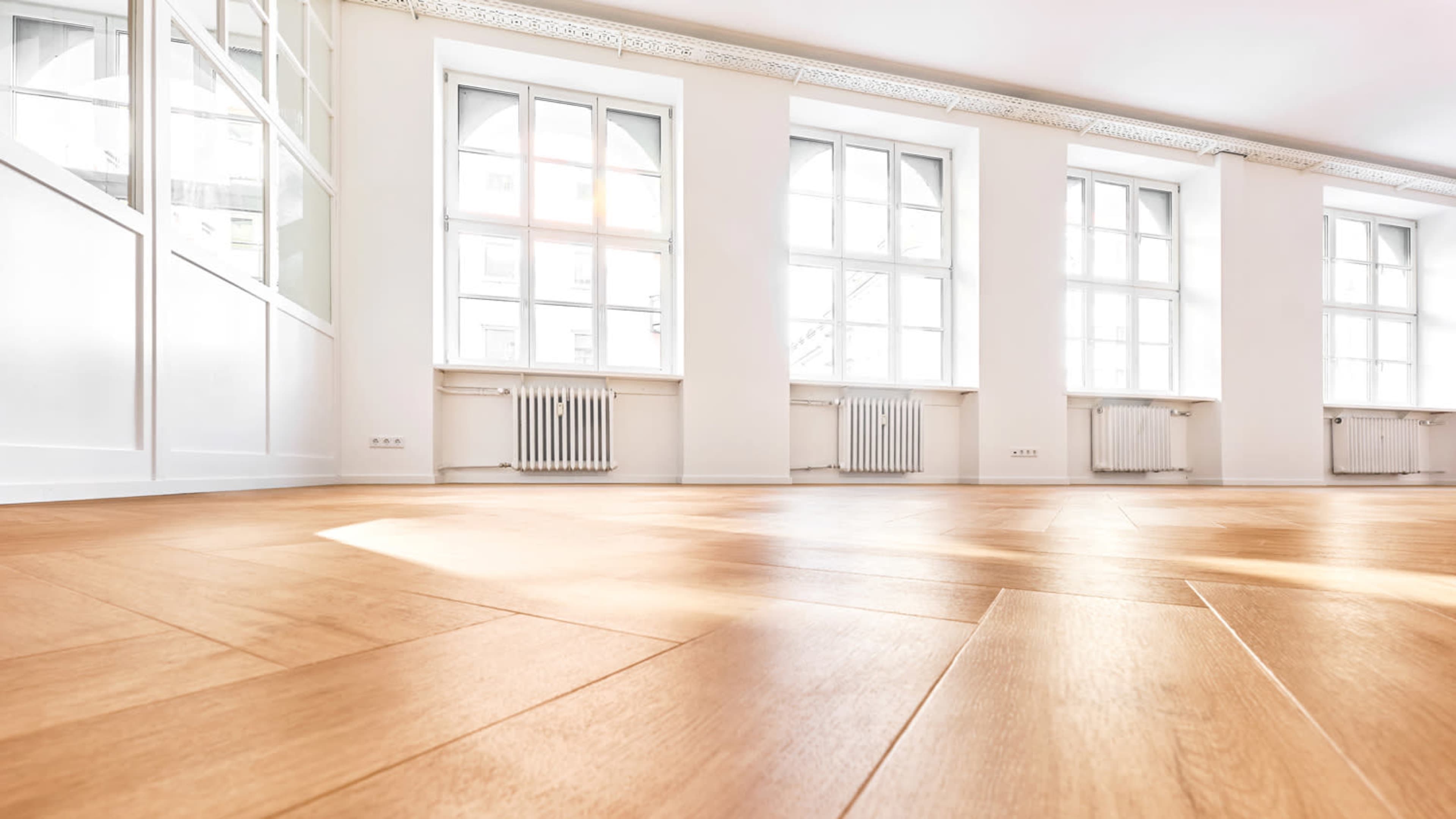 The image shows a spacious room with wooden flooring, large windows, and white walls.