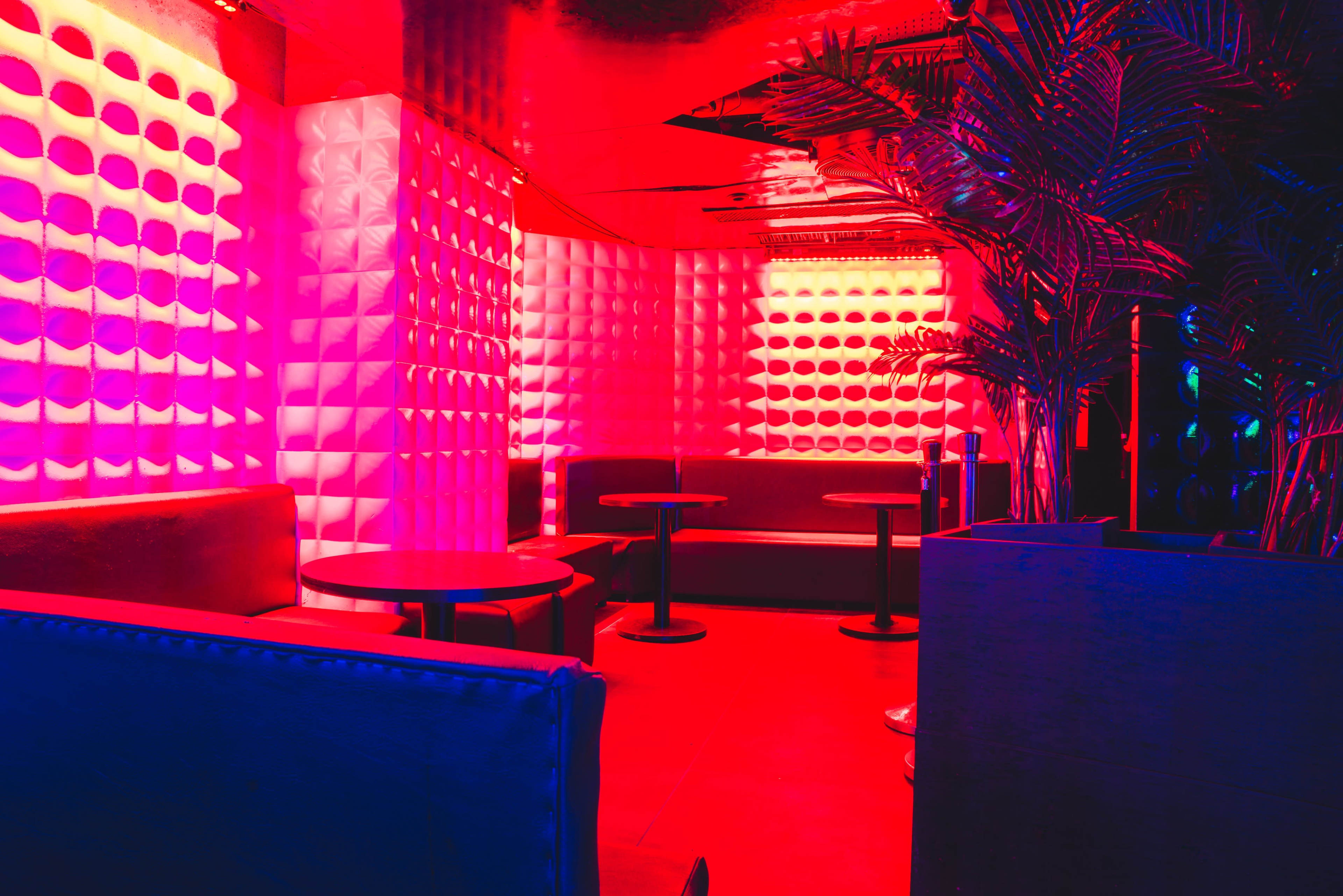 A dimly lit lounge features illuminated red and blue walls, with plush seating and small round tables.