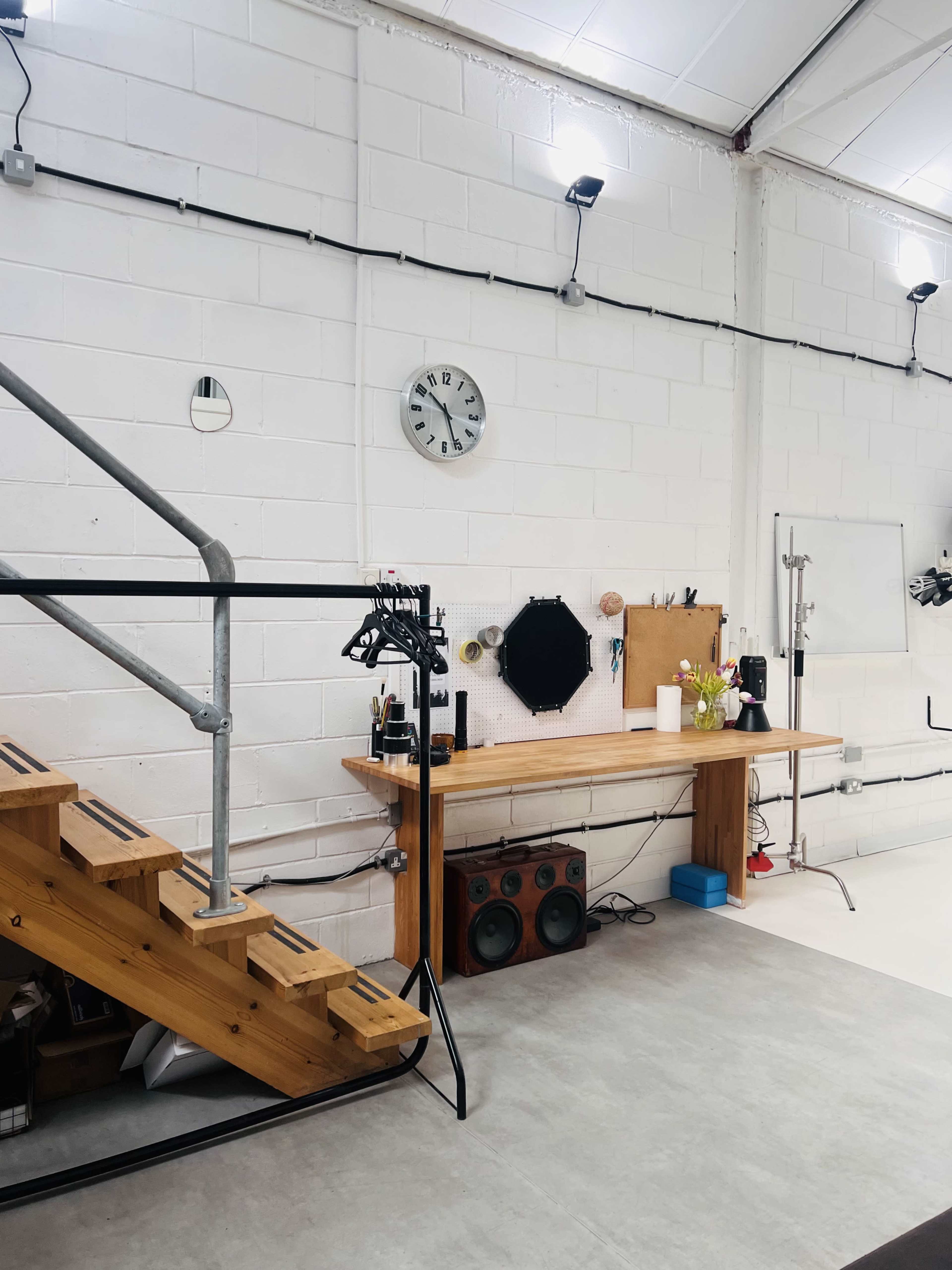 The image shows a well-lit industrial space featuring wooden stairs, a clock on the wall, a mirrored surface, and a workbench with various items arranged on it.