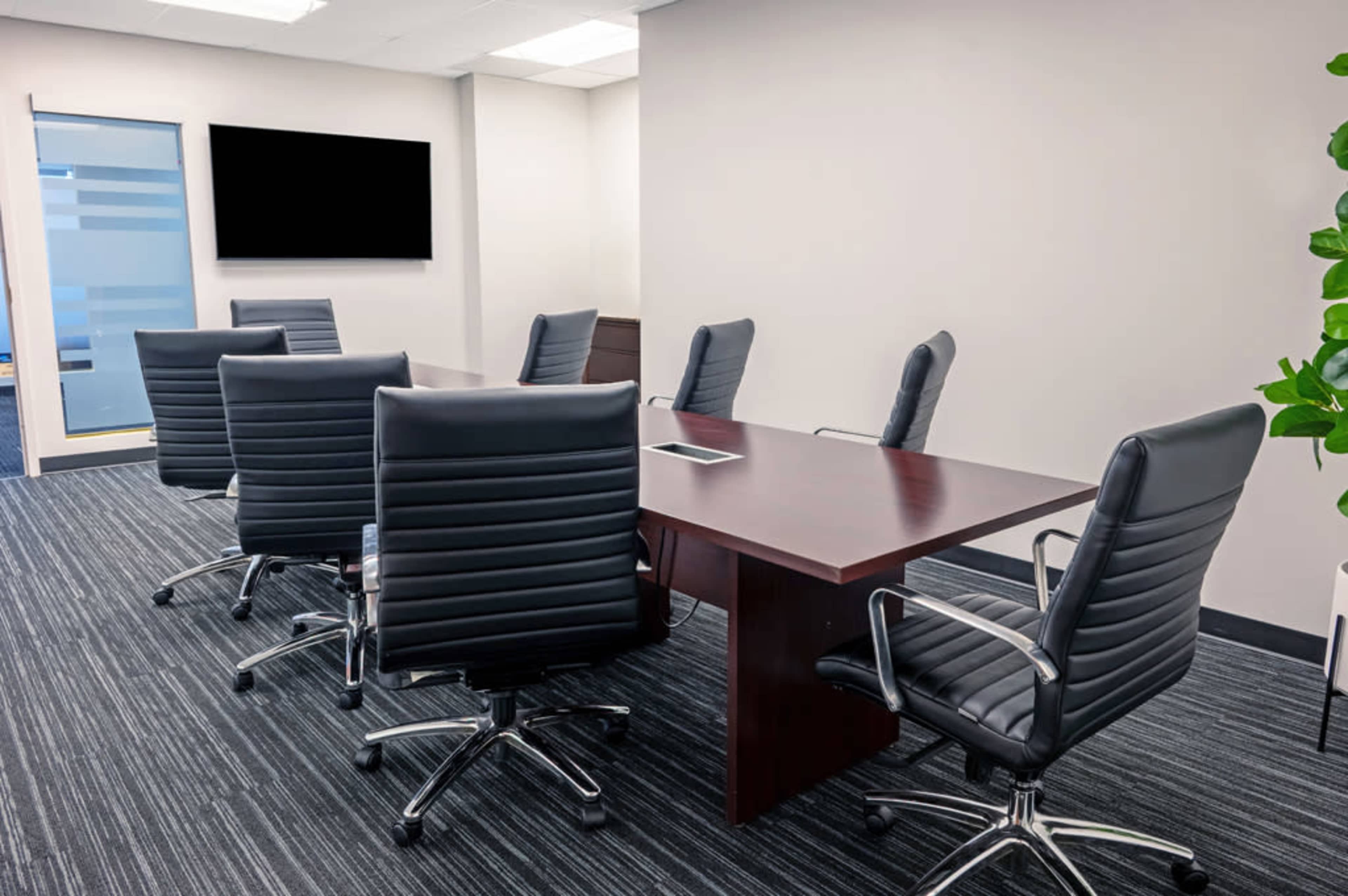 The image shows a modern conference room featuring a large brown table surrounded by black leather rolling chairs, with a television mounted on the wall.