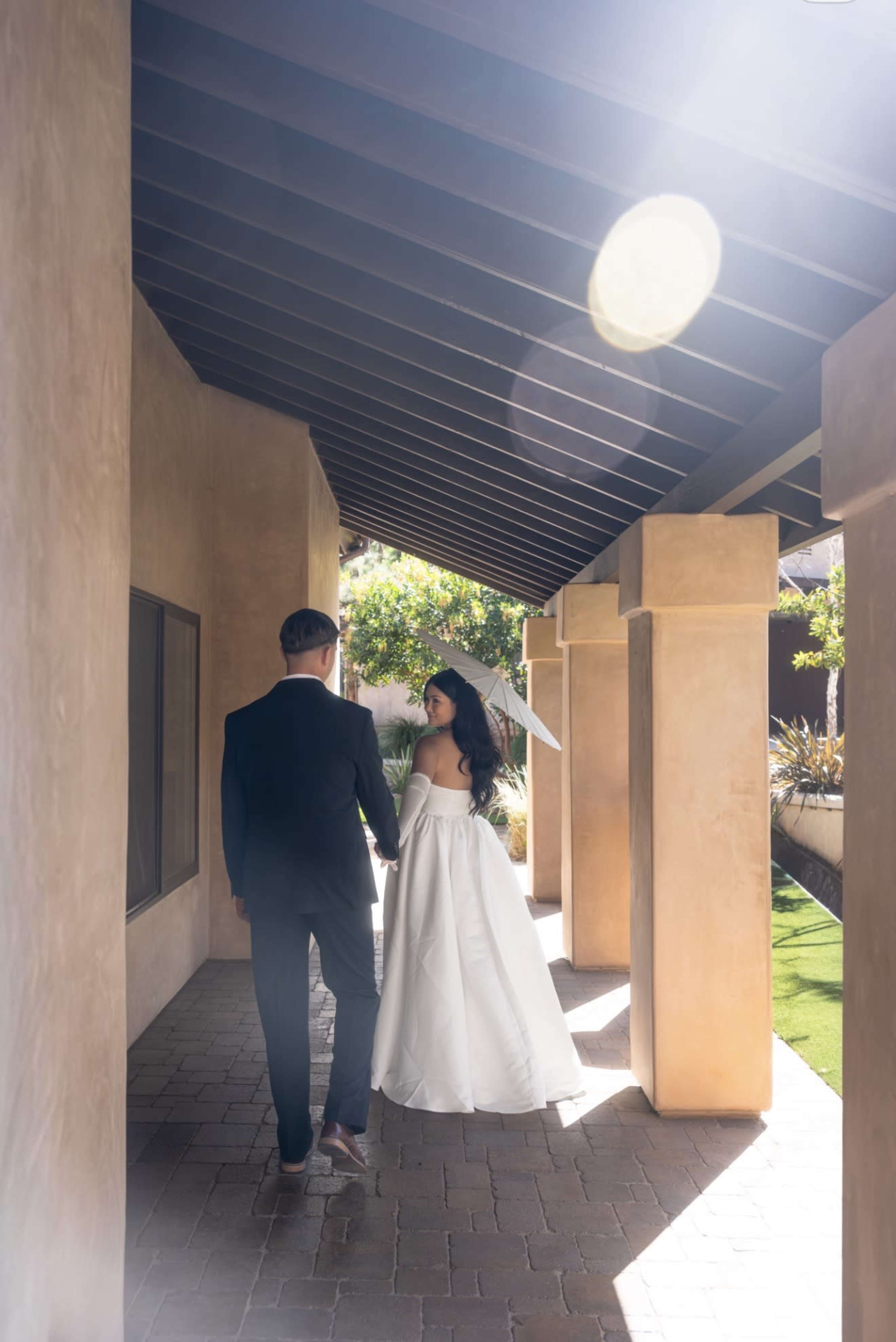 A couple walks hand-in-hand under a covered walkway, with the bride wearing a white gown and veil.
