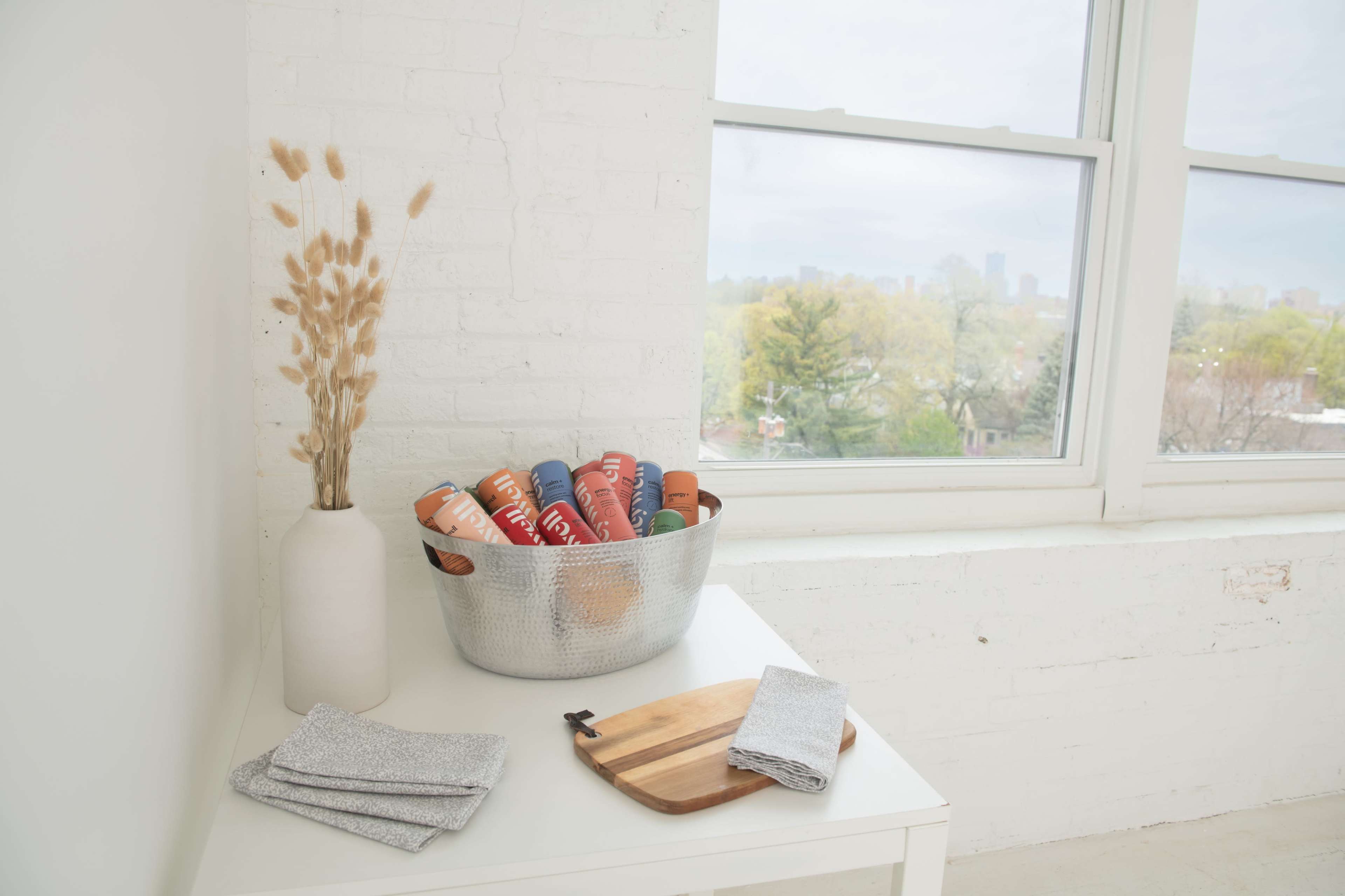 A silver bowl filled with colorful bottles sits on a white table next to a vase of dried plants and a window looking out at a green landscape.