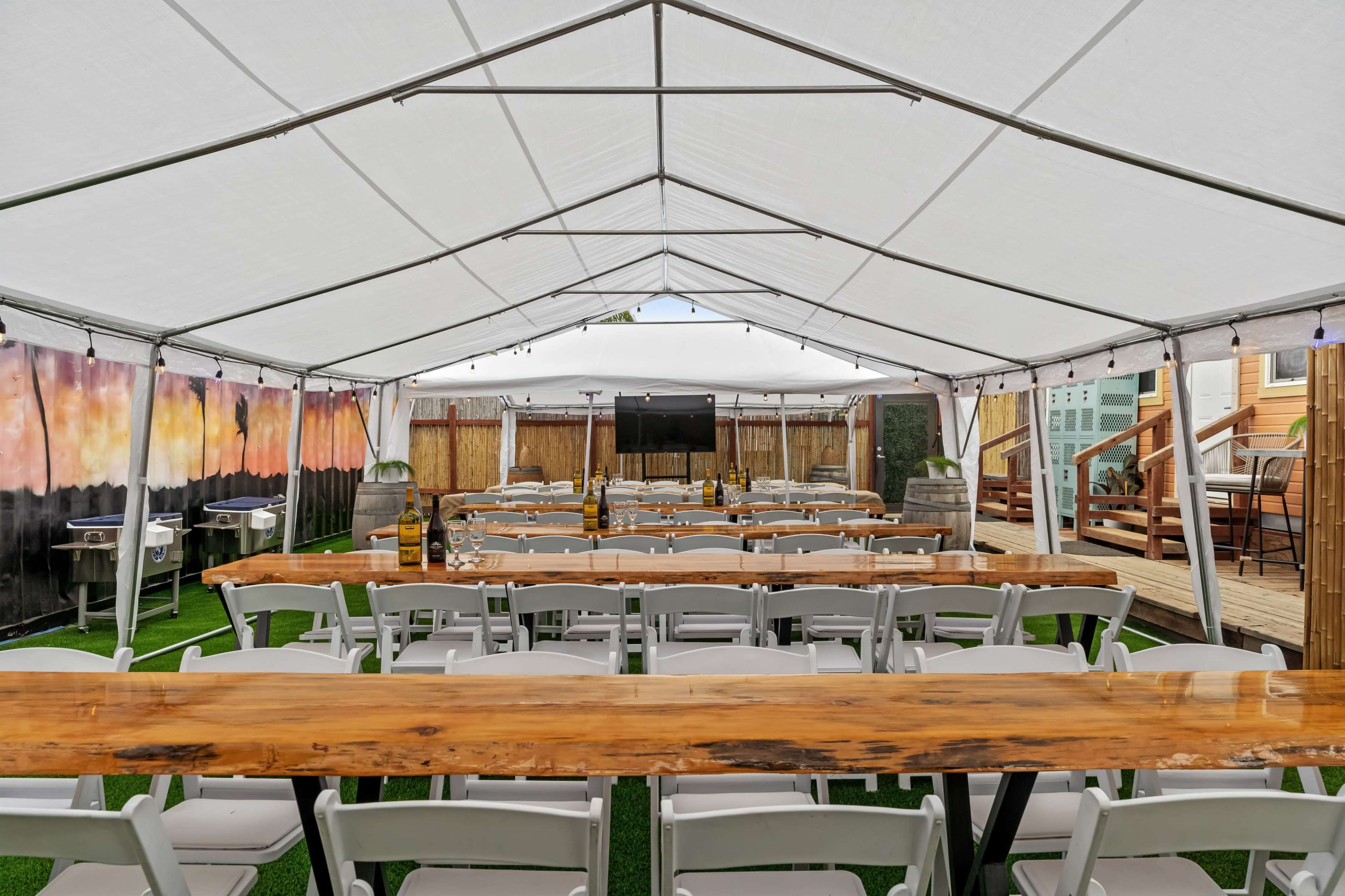 A large tent is set up with rows of white chairs facing a black screen, surrounded by wooden tables and a decorated backdrop.